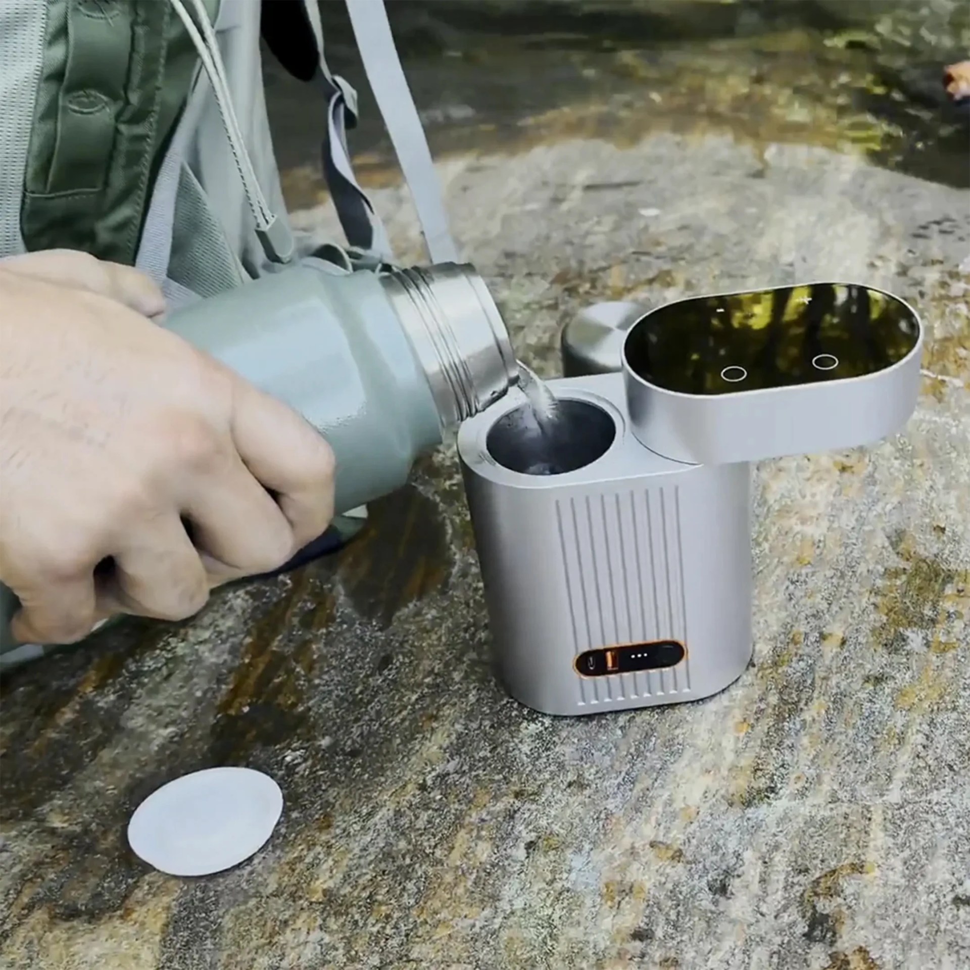 A hand is pouring water from a gray thermos into a small, silver, rectangular device with a black top and two circular touch buttons. The device is placed on a textured stone surface next to a green backpack. The device has a small display with orange indicators on its front.