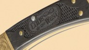 Close-up of a black knife handle with textured grip and brass rivets. The handle features an embossed logo that reads "COLONIAL IN THE WEST." The knife also has a brass bolster with decorative engraving.