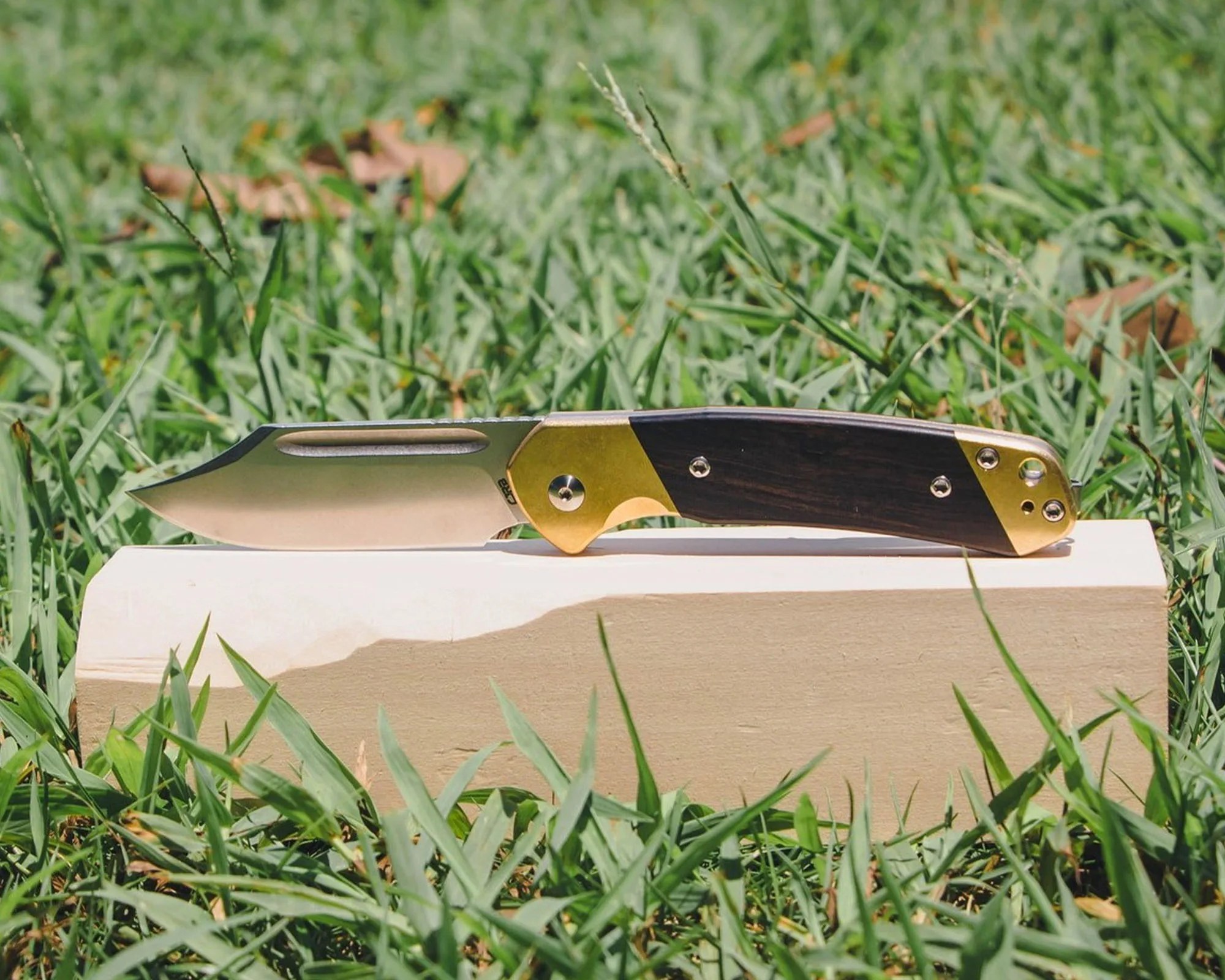 Folding knife with a polished silver blade and a handle featuring dark wood and brass accents, resting on a light-colored rectangular block placed on grass.