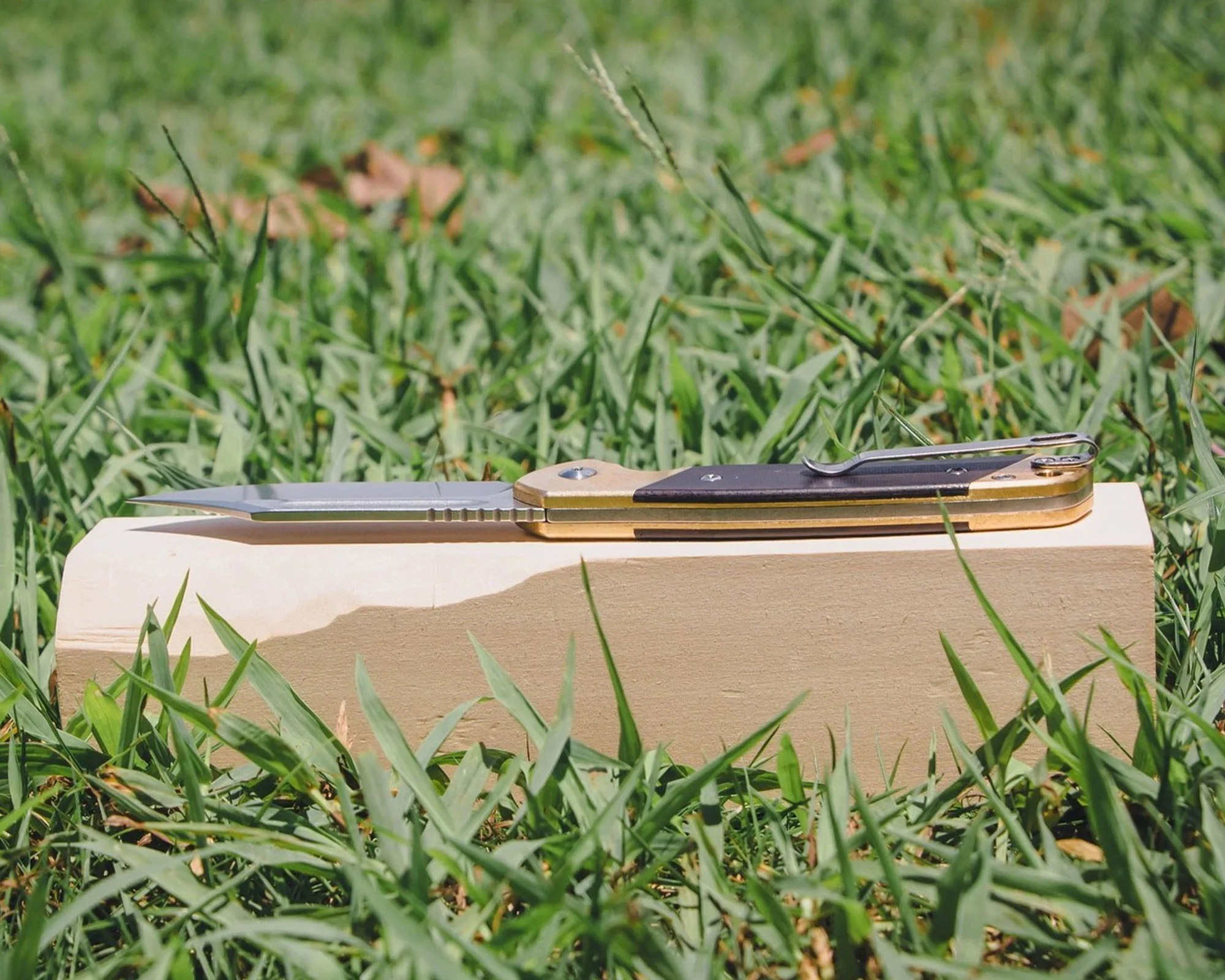 A folding knife with a partially serrated stainless steel blade and a handle featuring brass and dark wood accents, resting on a light-colored wooden block placed on green grass.