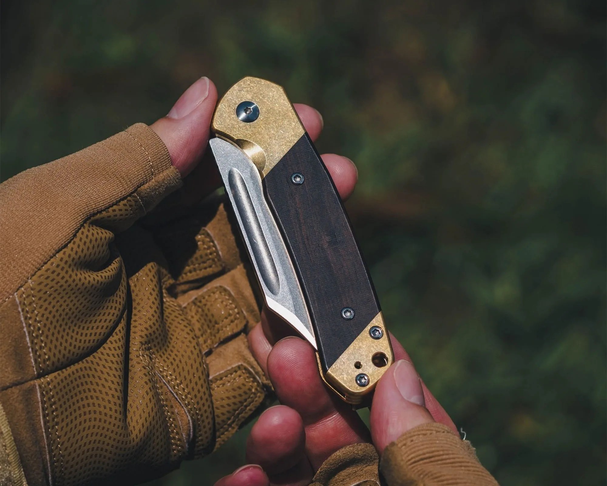 A hand wearing a brown tactical glove holding a closed folding knife with a brass and dark wood handle and a partially visible silver blade. The background is blurred greenery.