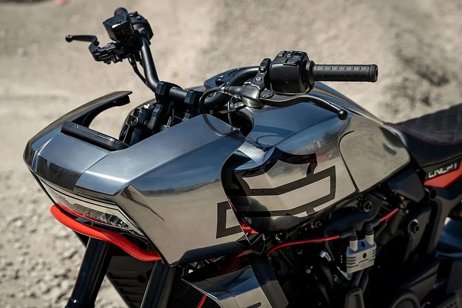 Close-up of a motorcycle's front section featuring a sleek, metallic silver fairing with a black geometric design. The handlebars are black with textured grips and control switches. A red accent bar is visible beneath the fairing, adding contrast to the design. The background is blurred, focusing attention on the motorcycle's details.