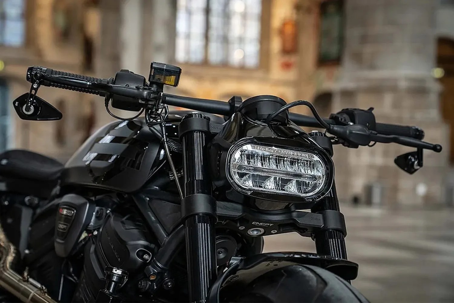 Close-up of the front section of a black Harley-Davidson motorcycle, highlighting the distinctive oval LED headlight, black handlebars with bar-end mirrors, and the front suspension forks. The background is softly blurred, showing an indoor setting with stone walls and large windows.