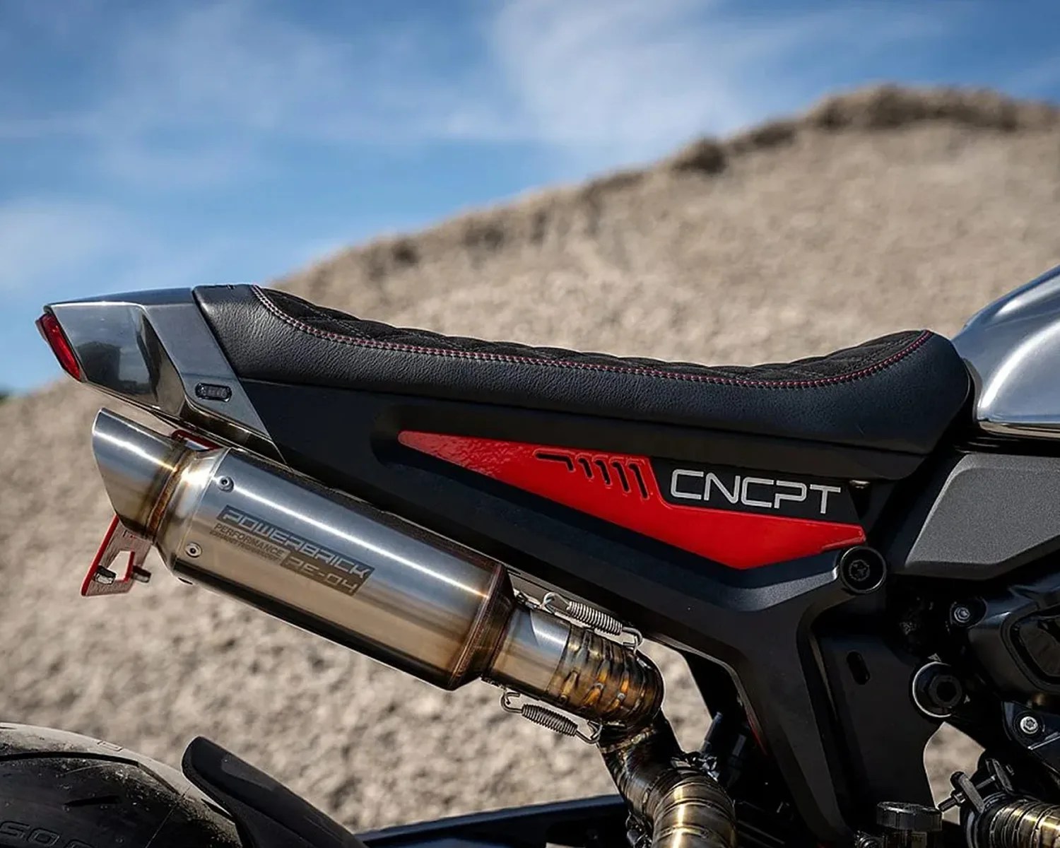 Close-up of a motorcycle's rear section featuring a black leather seat with red stitching, a red panel with the text "CNCPT," and a metallic exhaust pipe labeled "POWERBRICK PERFORMANCE PC-04." The background is blurred, showing a sandy or gravelly terrain under a blue sky.