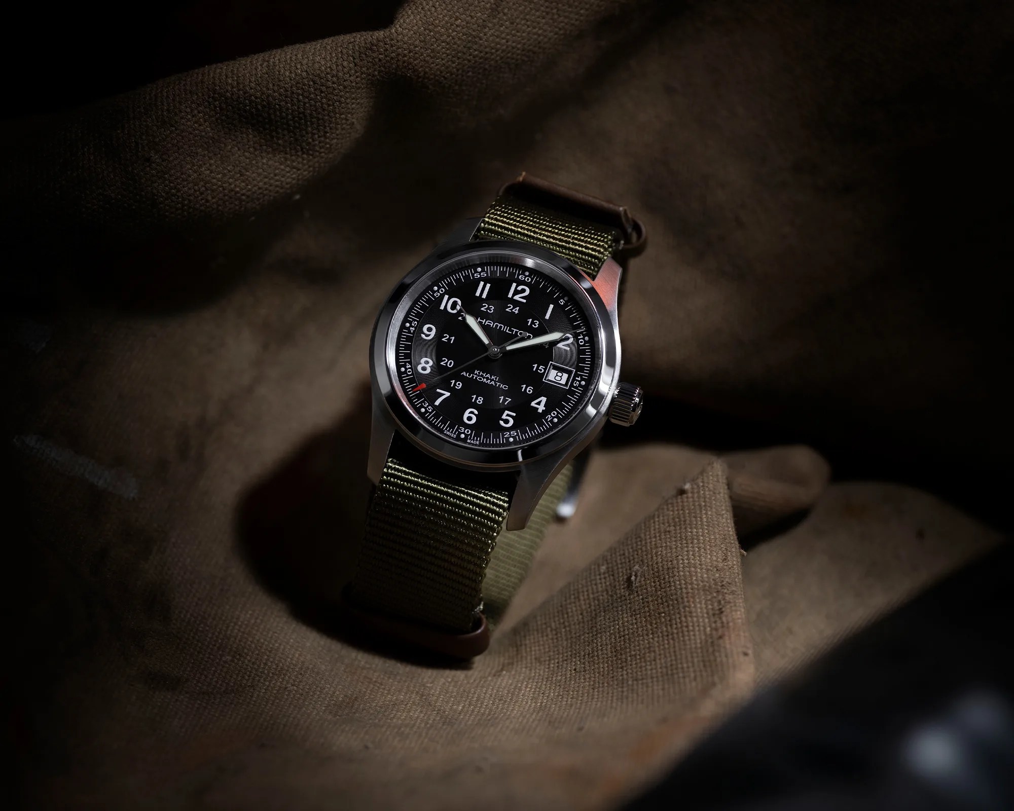 Hamilton Khaki Automatic wristwatch with a black dial, white numerals, and a date window at 3 o'clock, featuring a green fabric strap, placed on a textured brown fabric background.