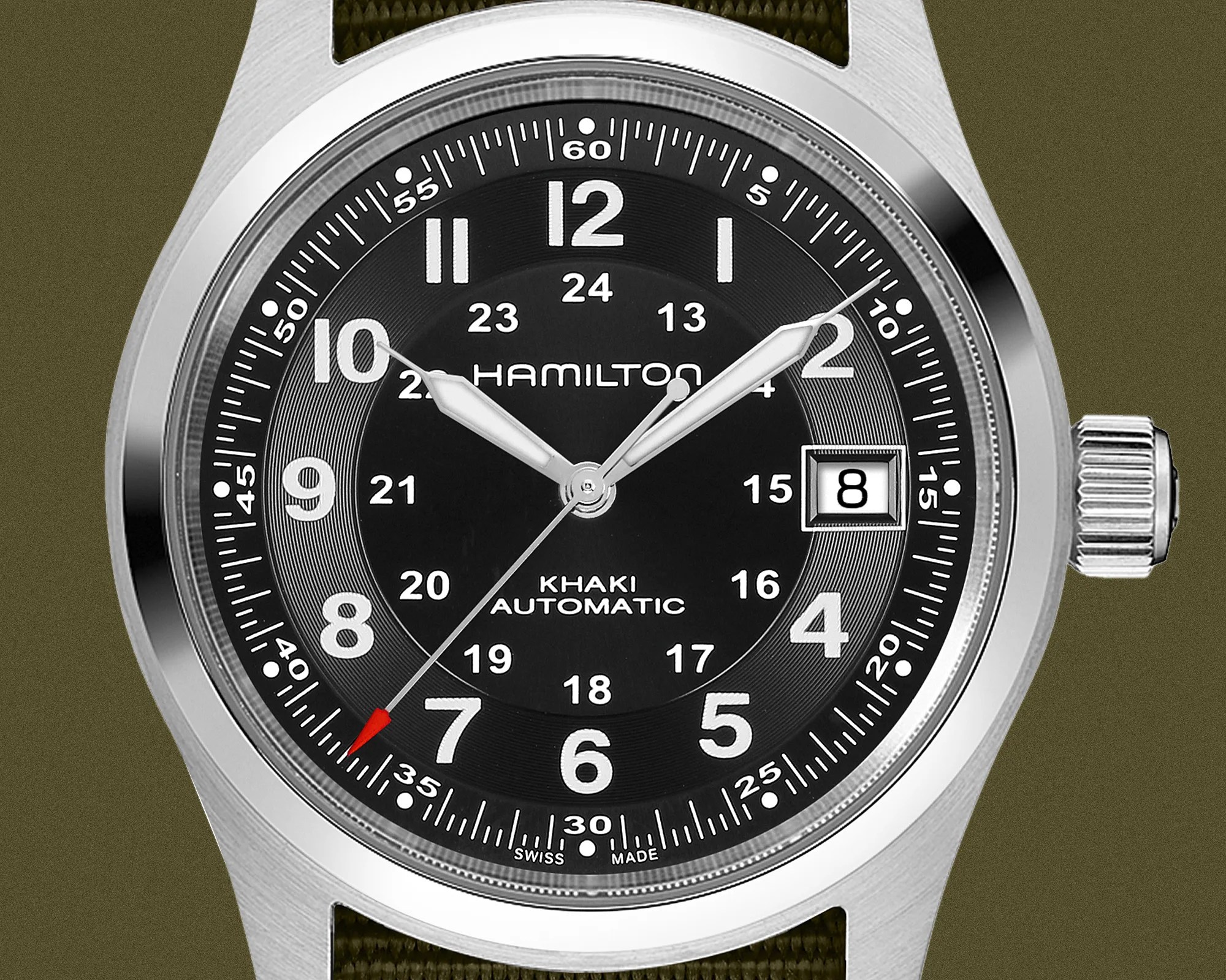 Hamilton Khaki Automatic wristwatch with a black dial featuring large white Arabic numerals for hours, smaller 24-hour military time numerals inside, and a date window at 3 o'clock. The watch has silver-tone hands with luminous fill, a red-tipped second hand, and a stainless steel case with a textured crown. The strap is olive green fabric.