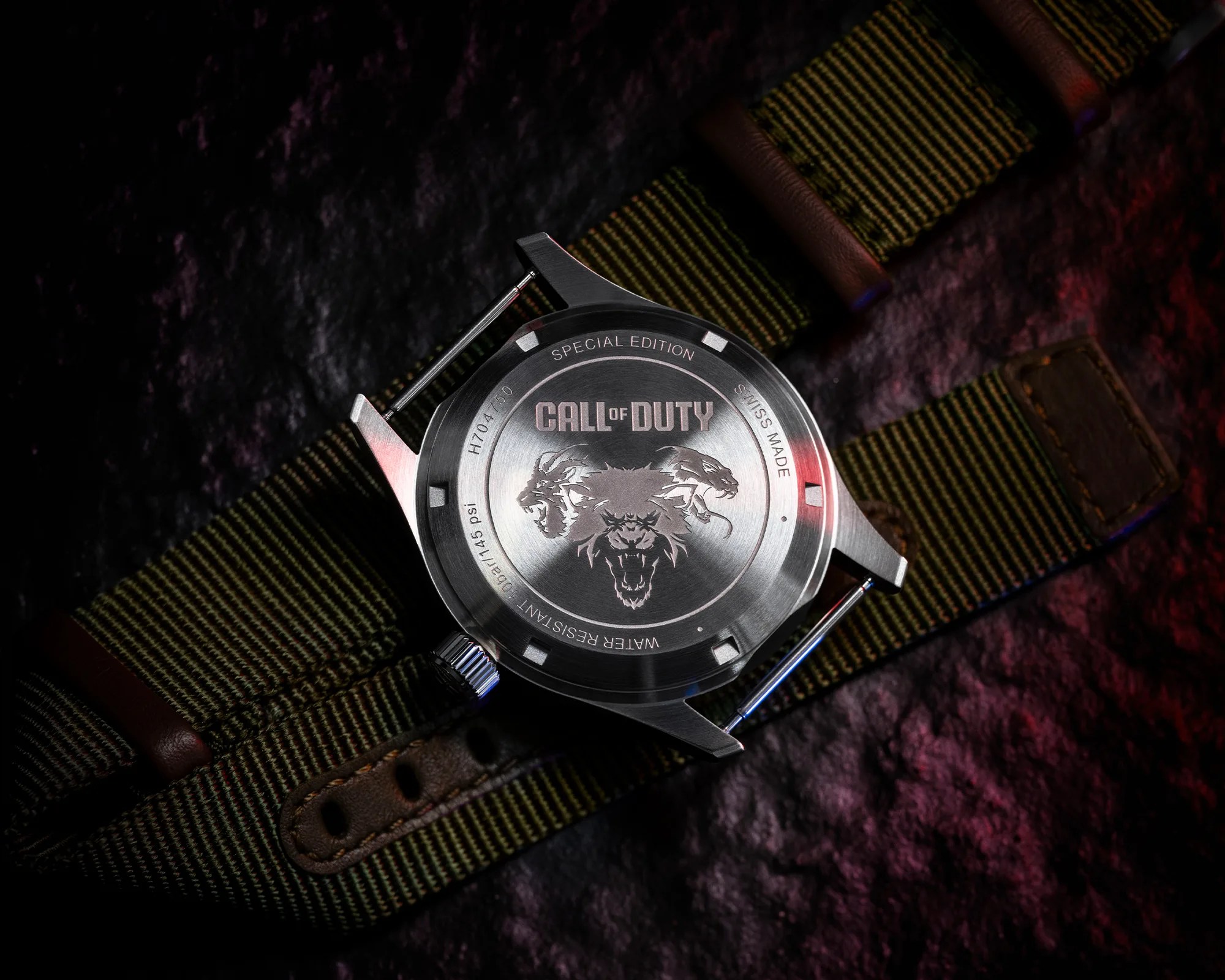 Back view of a special edition Call of Duty wristwatch with a stainless steel case. The case back features an engraved design of three roaring animal heads and the text "CALL OF DUTY," "SPECIAL EDITION," "SWISS MADE," and "WATER RESISTANT 10BAR/145 psi." The watch has a green and brown striped fabric strap with leather accents. The background is dark with subtle red and blue lighting.