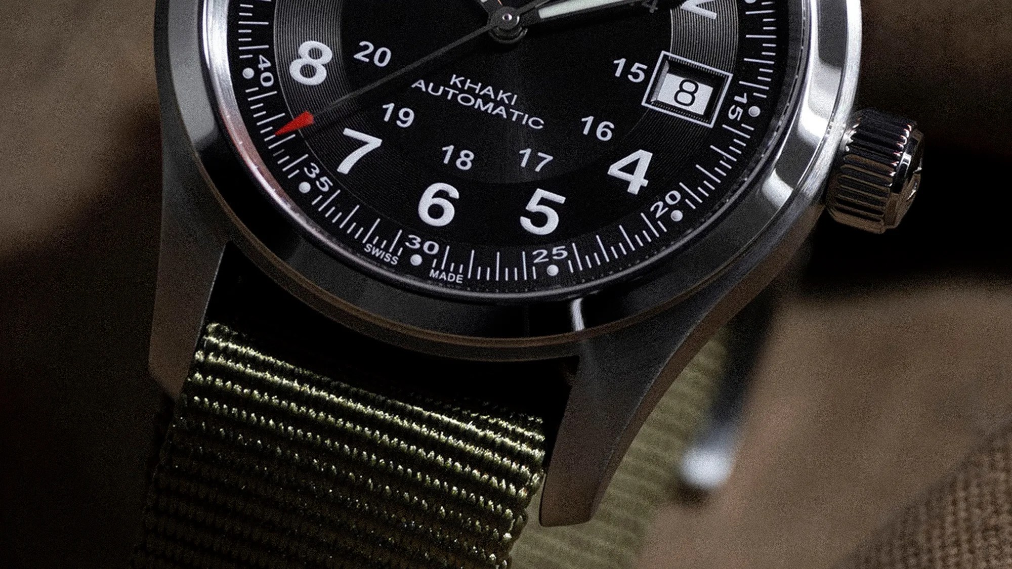Close-up of a wristwatch with a black dial featuring white numerals and markings, a date window showing the number 8, and the text "KHAKI AUTOMATIC" and "SWISS MADE." The watch has a silver bezel and a textured green fabric strap. The second hand is red.