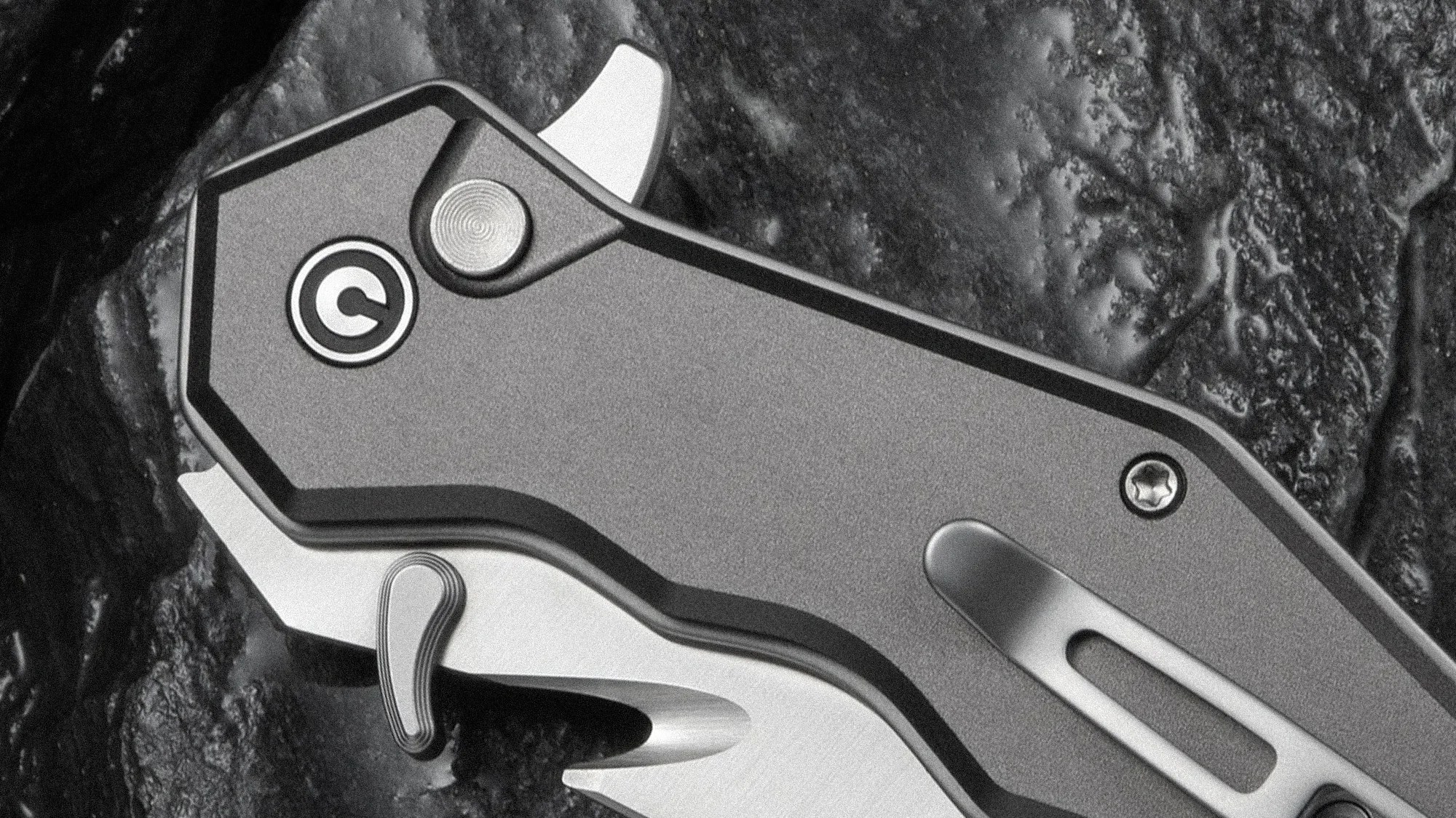 Close-up of a folding knife handle with a textured grip, a circular button, a clip, and a partially visible blade. The handle has a matte finish and a logo resembling a "C" inside a circle near the button. The background is a rough, dark surface.