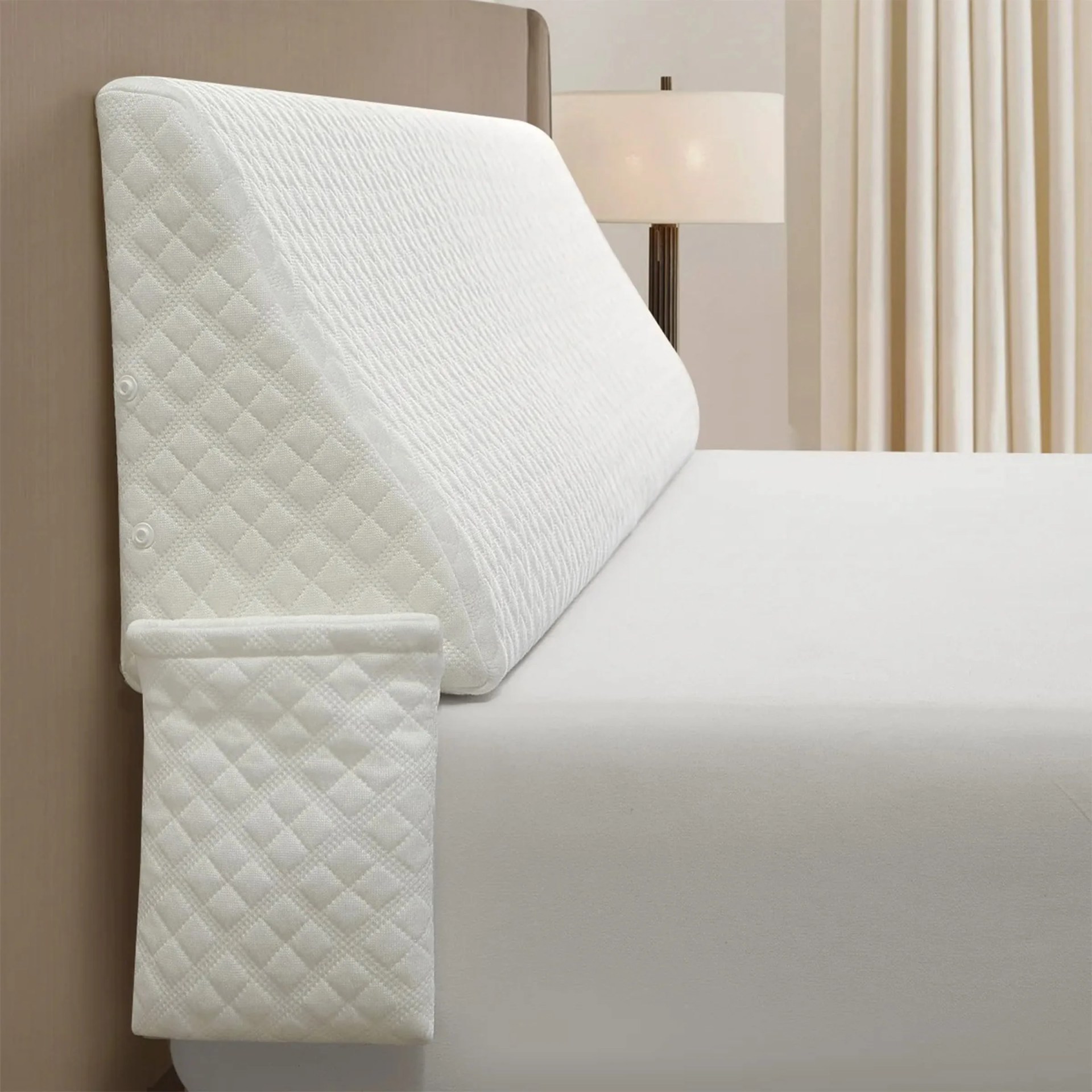 White quilted wedge pillow with a matching quilted pocket attached to the side, placed on a bed with a beige headboard, next to a lamp and cream-colored curtains.