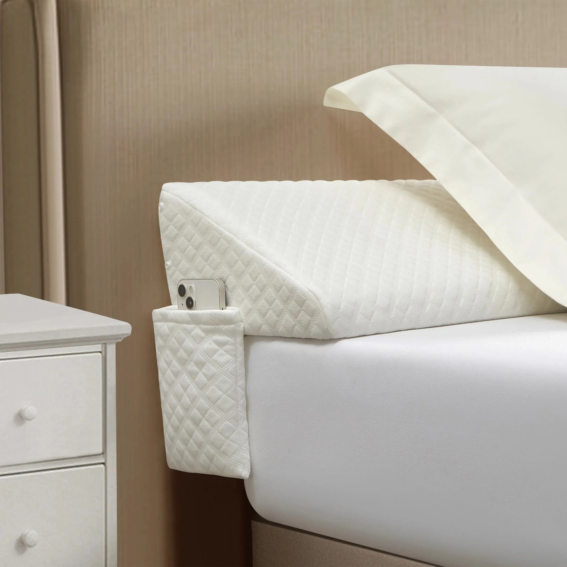 A white wedge pillow with a quilted texture is placed on a bed, partially covered by a white pillowcase. The pillow has a built-in pocket on the side, holding a smartphone. The bed has a white fitted sheet, and a white nightstand with drawers is visible next to the bed. The background features a beige wall.