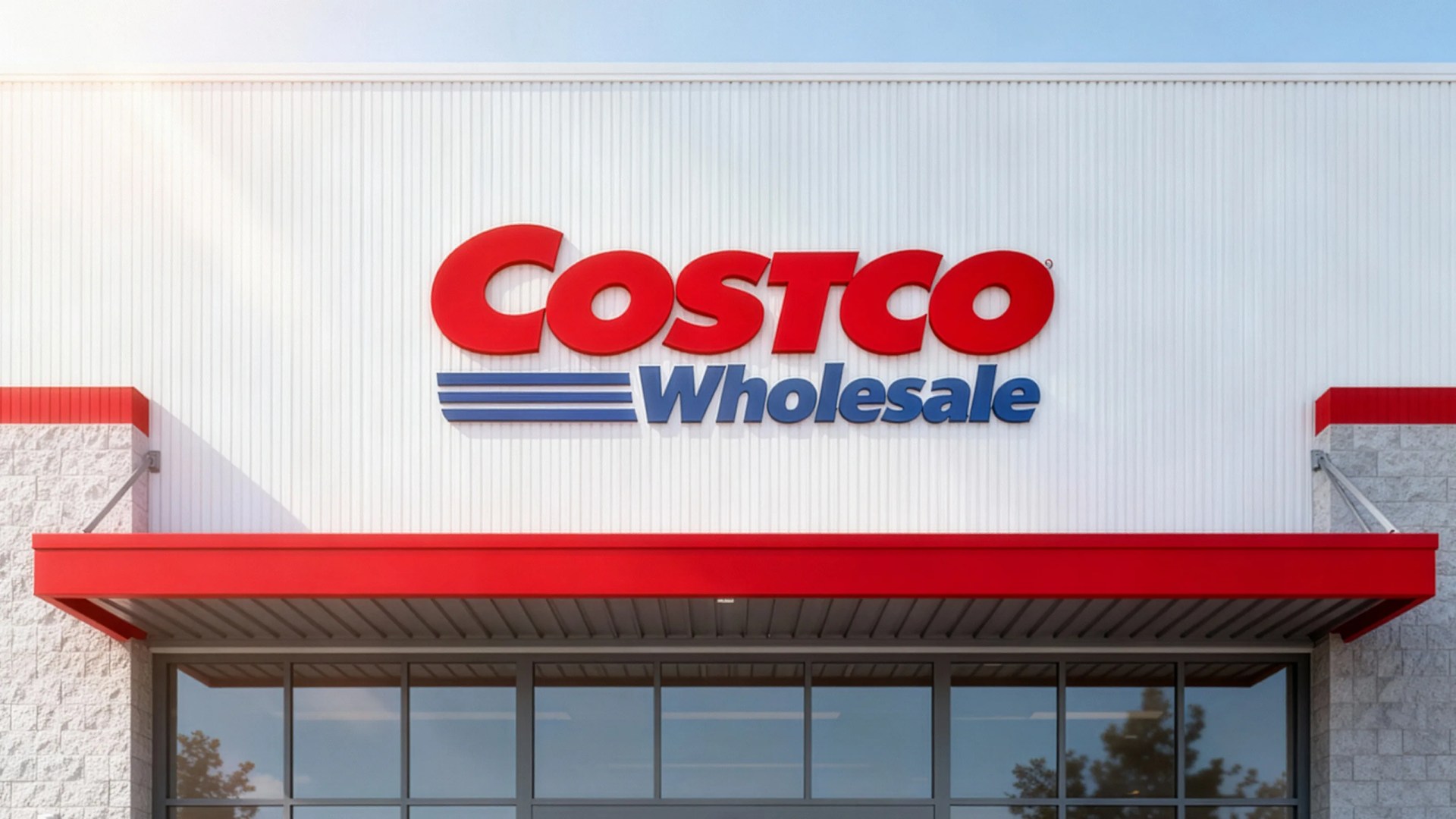 Costco Wholesale store entrance with a white exterior wall, red and blue logo, and a red awning above glass doors.