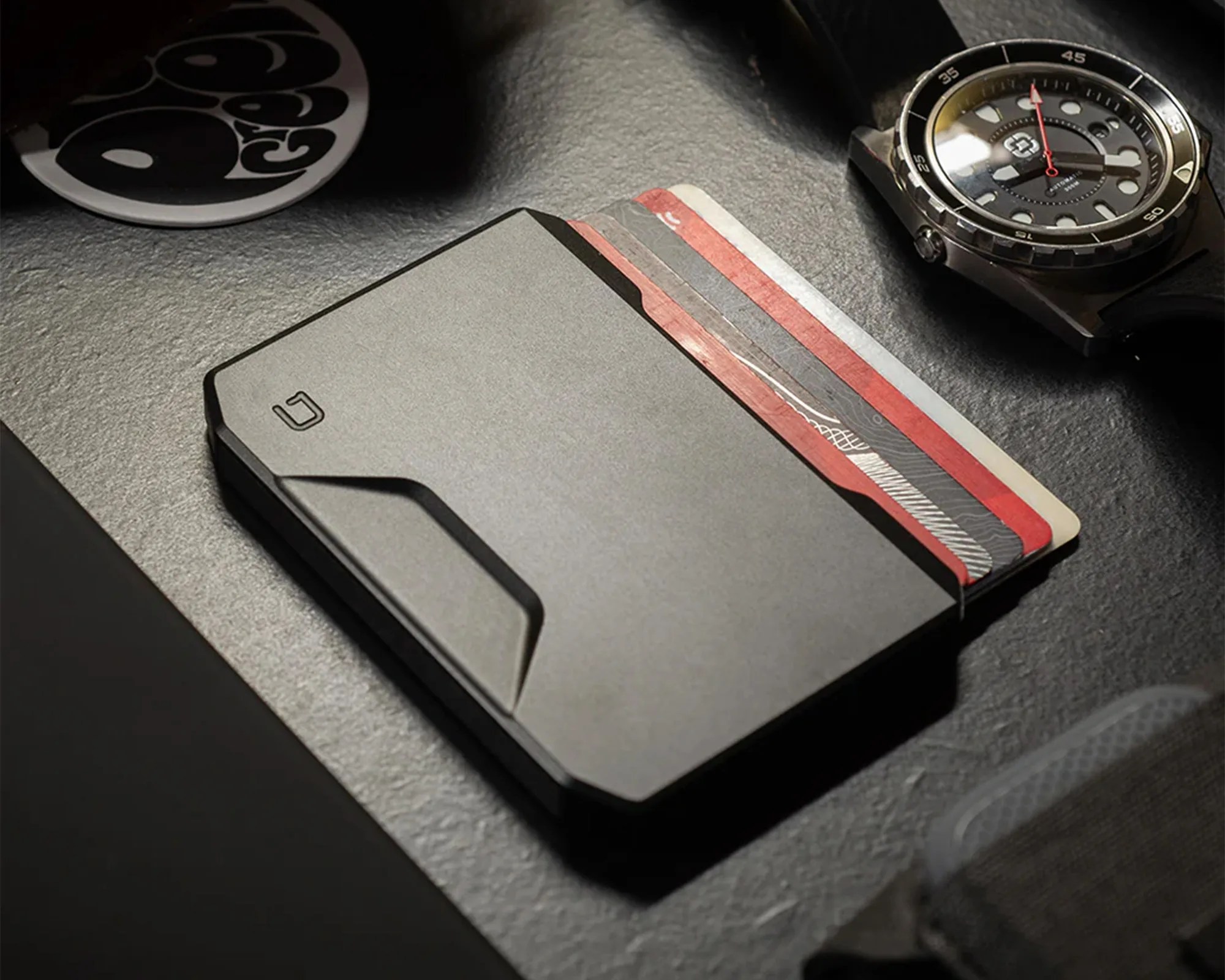 A sleek, black minimalist wallet holding several cards, placed on a textured dark surface. Nearby, there is a black wristwatch with a round face and a black strap, and a round black and white sticker partially visible in the upper left corner.