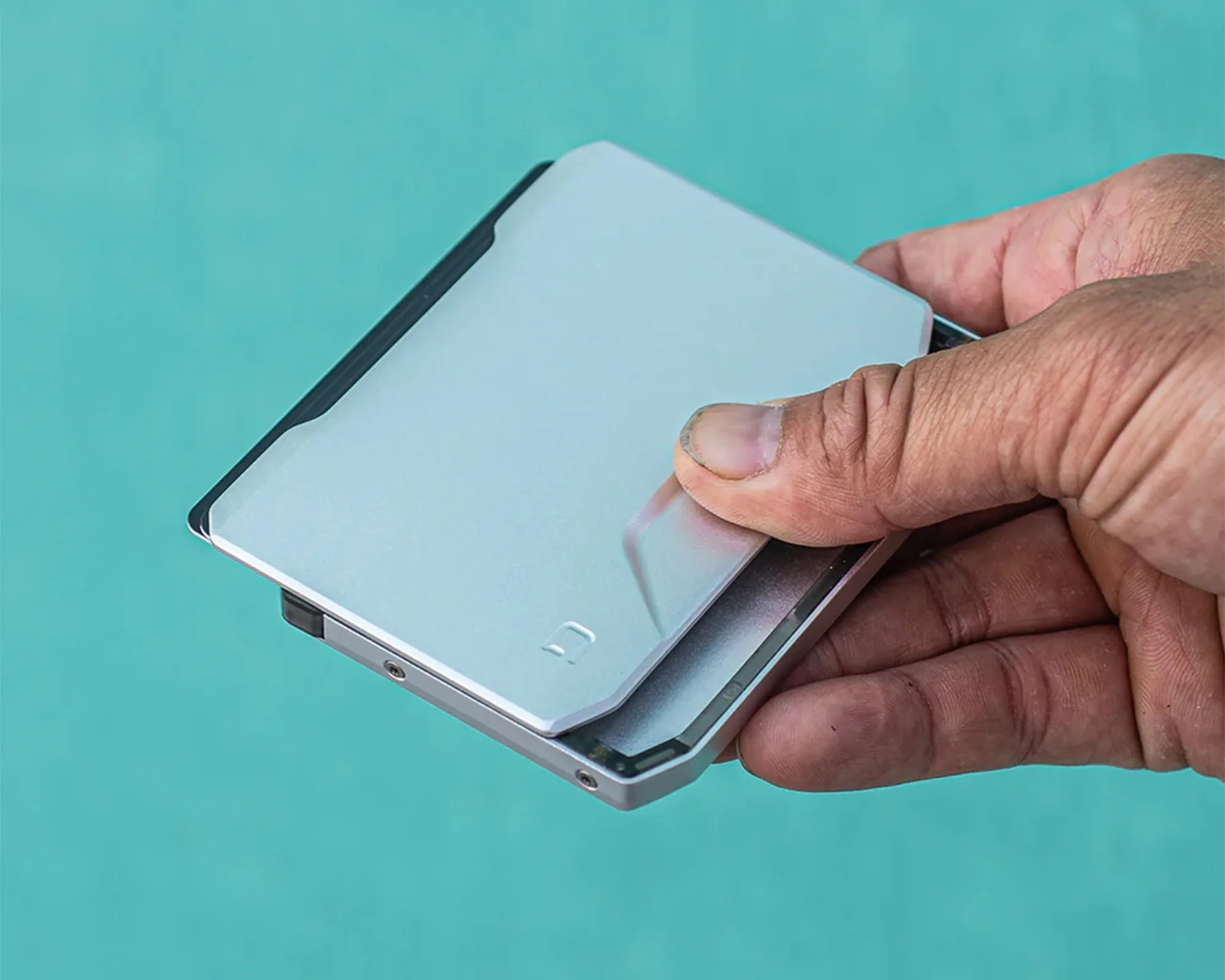 A hand holding a slim, metallic silver wallet with a minimalist design, featuring a small embossed logo on the front. The wallet has a sleek, modern appearance with clean lines and a compact form.