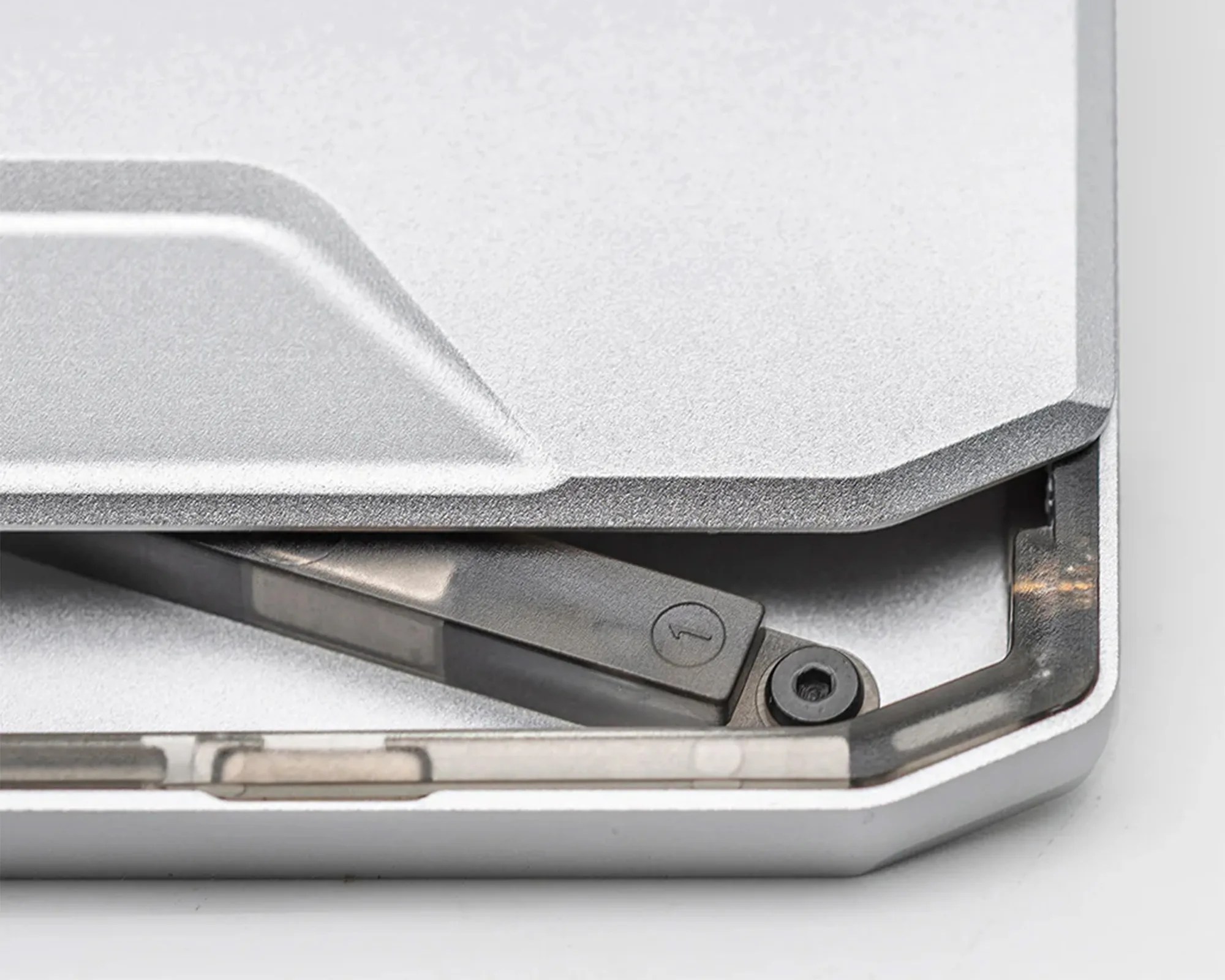 Close-up of a metallic hinge mechanism partially enclosed within a silver, textured casing, showing a black pivot arm with a circular marking and a screw, likely part of a device or machinery.