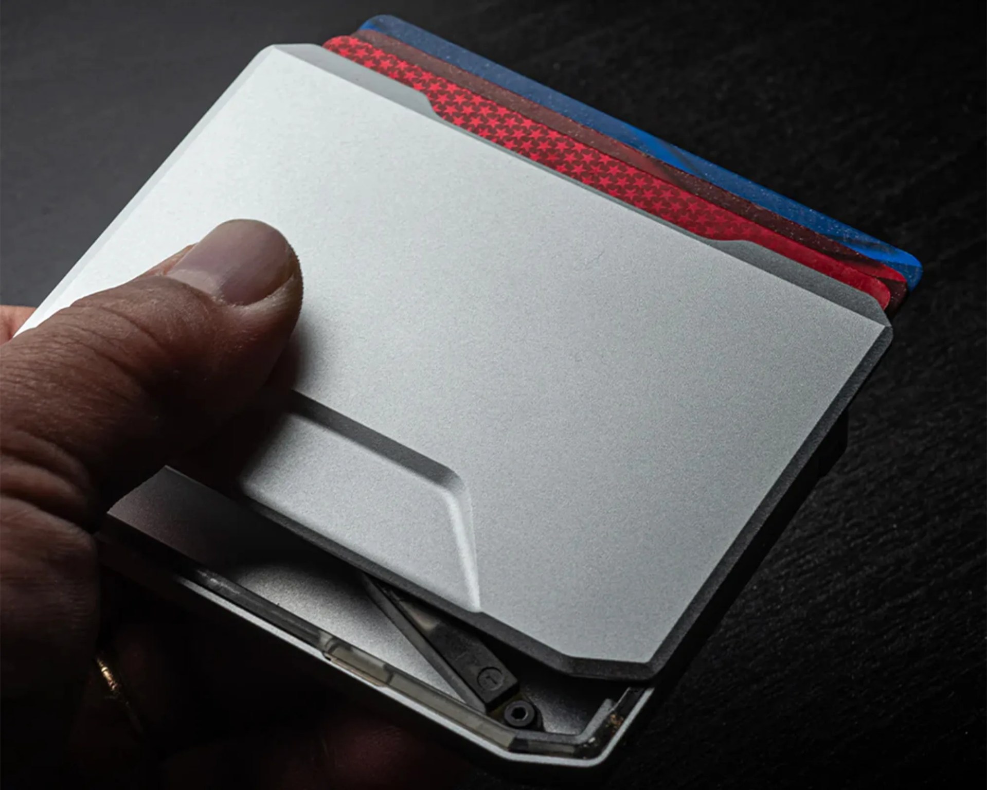 A hand holding a slim, metallic cardholder wallet with three visible cards inside, colored blue, red with stars, and brown. The wallet has a minimalist design with a small indentation for easy card access.