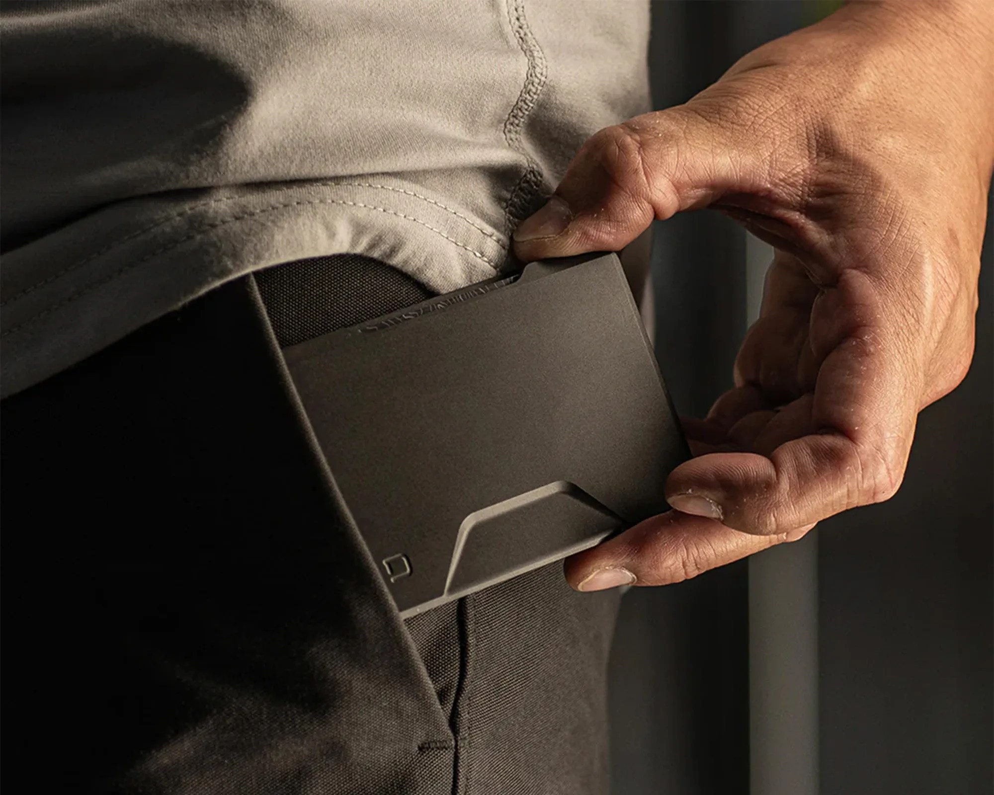 A hand is placing a slim, black wallet into the front pocket of dark pants. The person is wearing a light gray shirt. The wallet has a minimalist design with a small, subtle logo in the bottom left corner.