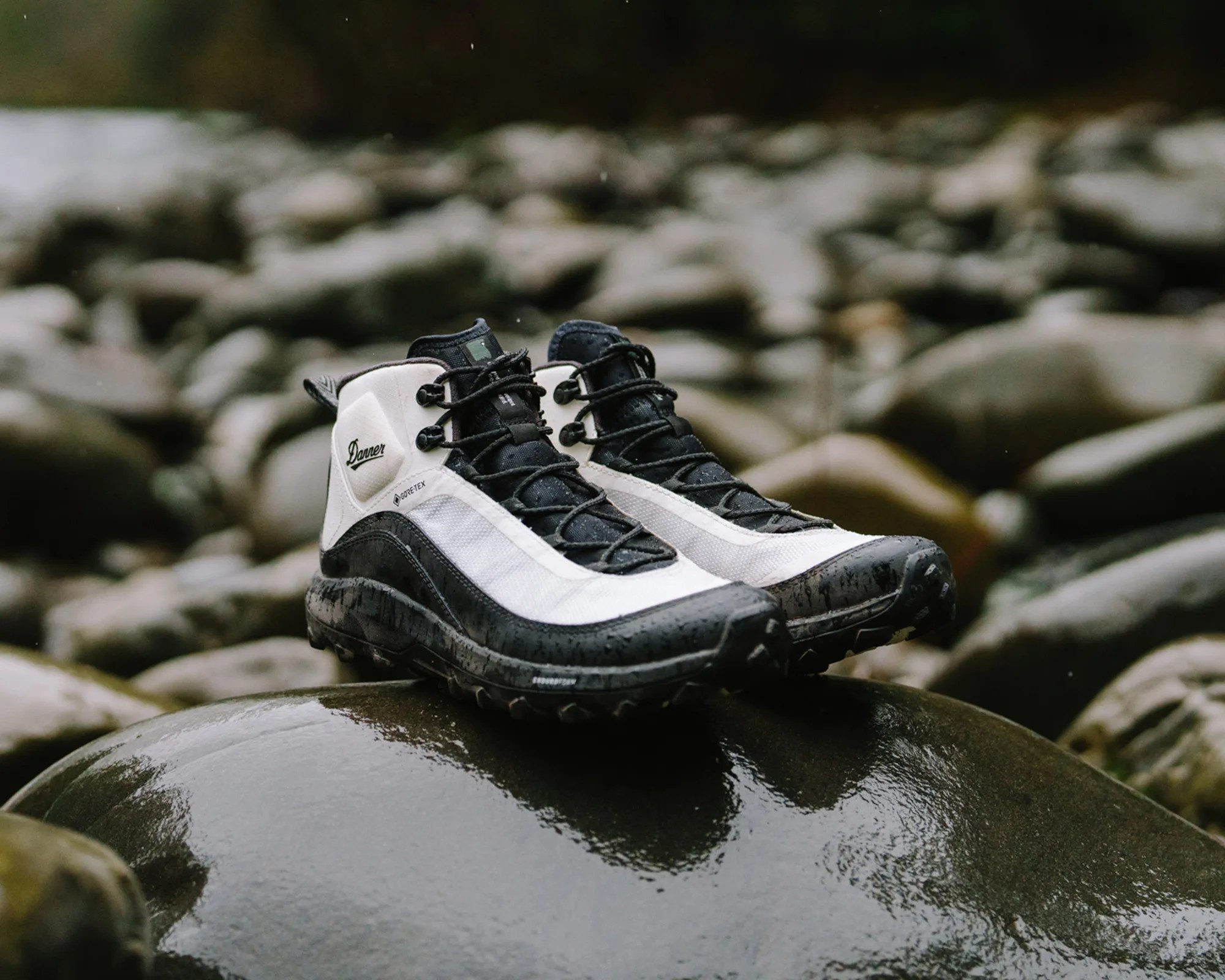 Pair of black and white Gore-Tex hiking boots with black laces, placed on a wet, smooth rock surrounded by other rocks. The boots have a rugged sole and the brand name "Danner" visible on the side.