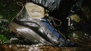 A close-up of a wet hiking boot with the brand name "Danner" and a "GORE-TEX" label, featuring a combination of black, blue, and tan materials. The boot is being worn, and water droplets are visible around it, suggesting use in a wet environment.