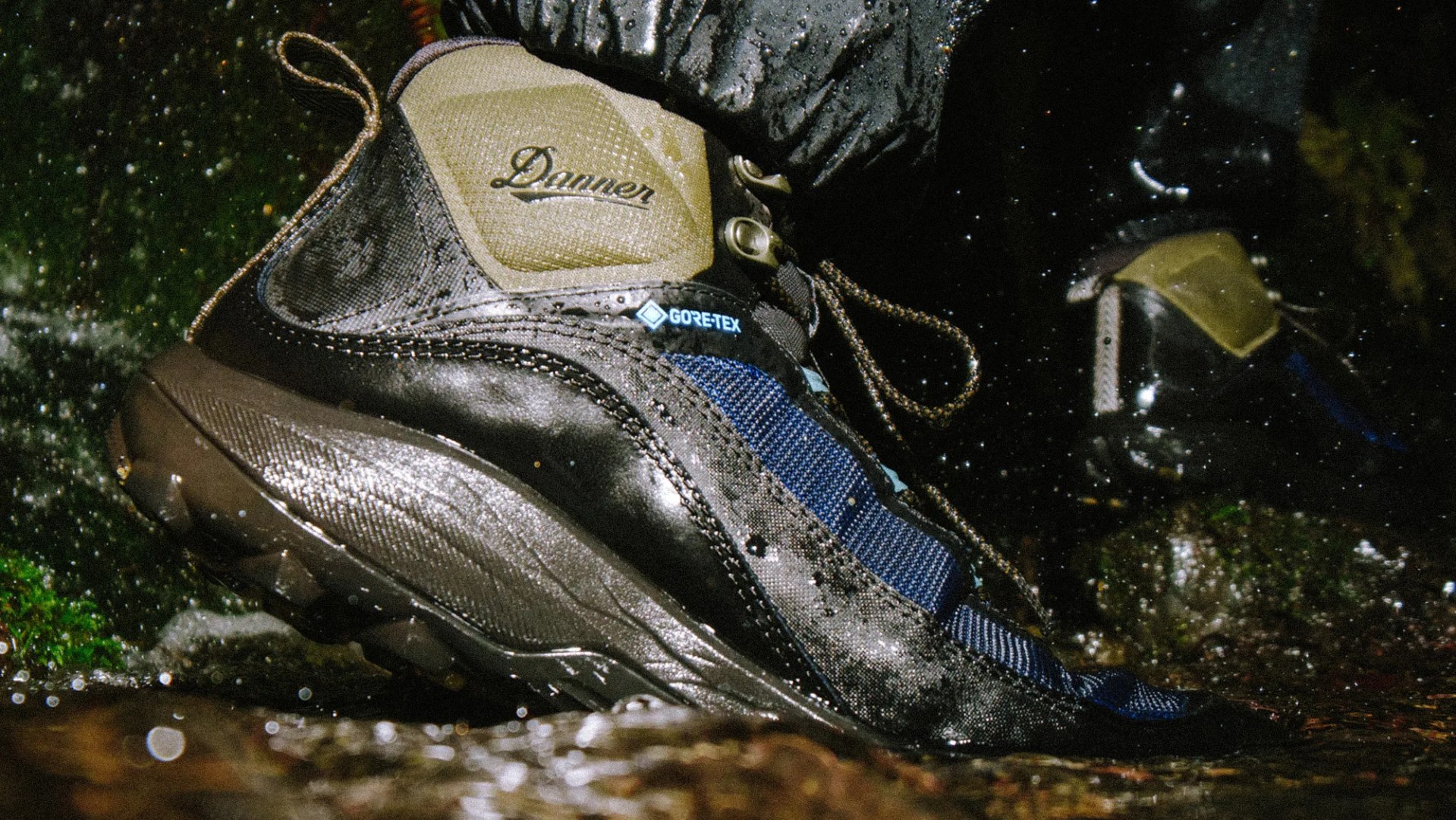 A close-up of a wet hiking boot with the brand name "Danner" and a "GORE-TEX" label, featuring a combination of black, blue, and tan materials. The boot is being worn, and water droplets are visible around it, suggesting use in a wet environment.