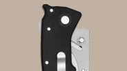 Close-up of a folding knife handle with a textured black grip, three visible rivets, a circular pivot screw, and a partially visible silver metal blade with a thumb stud. The background is a plain beige color.
