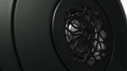 Close-up of a black speaker cone with a textured surround and a decorative grille featuring an abstract, organic pattern. The speaker is set within a matte black circular frame.