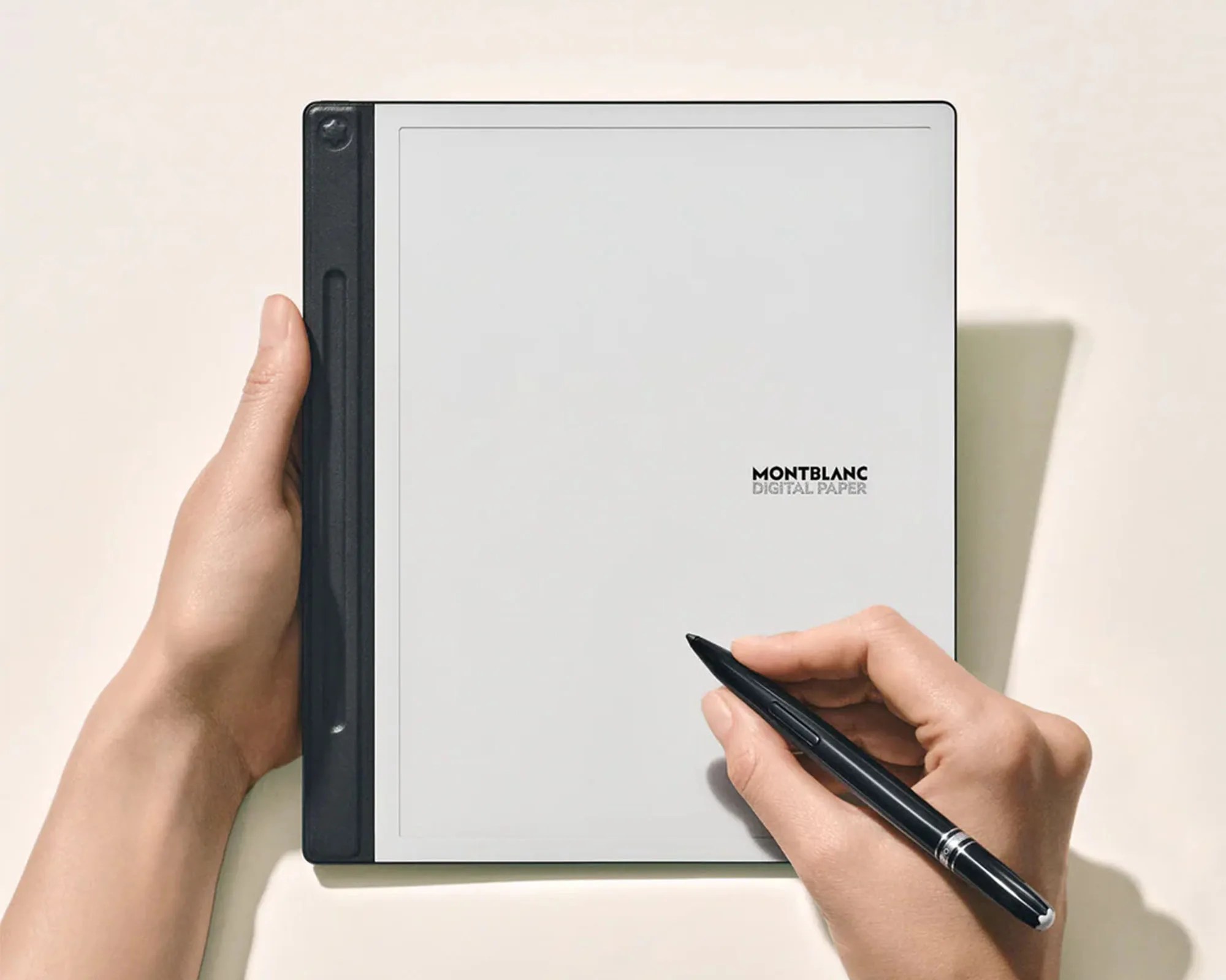A person holding a Montblanc Digital Paper tablet with one hand while writing on it with a black Montblanc pen in the other hand. The tablet has a large, light gray screen with the Montblanc logo and "DIGITAL PAPER" text near the center. The device has a black spine on the left side.