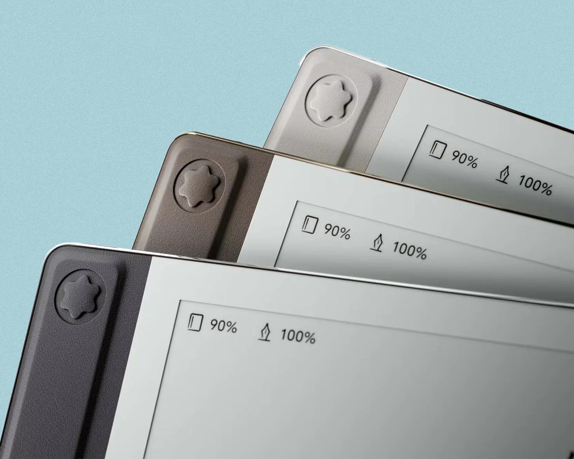 Three e-readers stacked diagonally, each showing a battery level of 90% and a pen battery level of 100%, with the devices featuring a textured star-shaped button on the top left corner in different shades of gray, beige, and dark gray against a light blue background.
