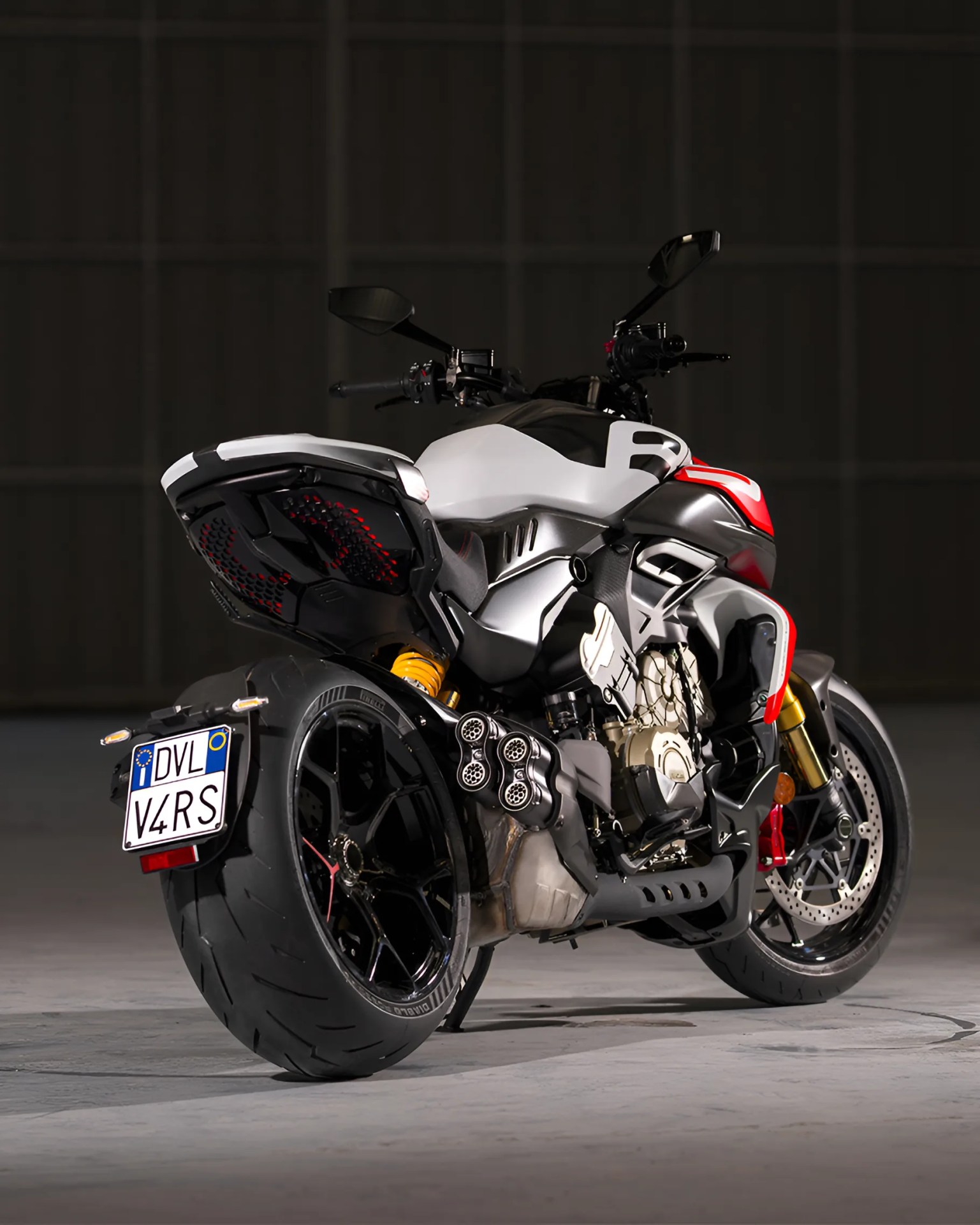 Sport motorcycle with a wide rear tire, black and white body with red accents, dual exhaust pipes on the right side, and a European-style license plate reading "DVL V4RS." The bike is photographed indoors against a dark background.