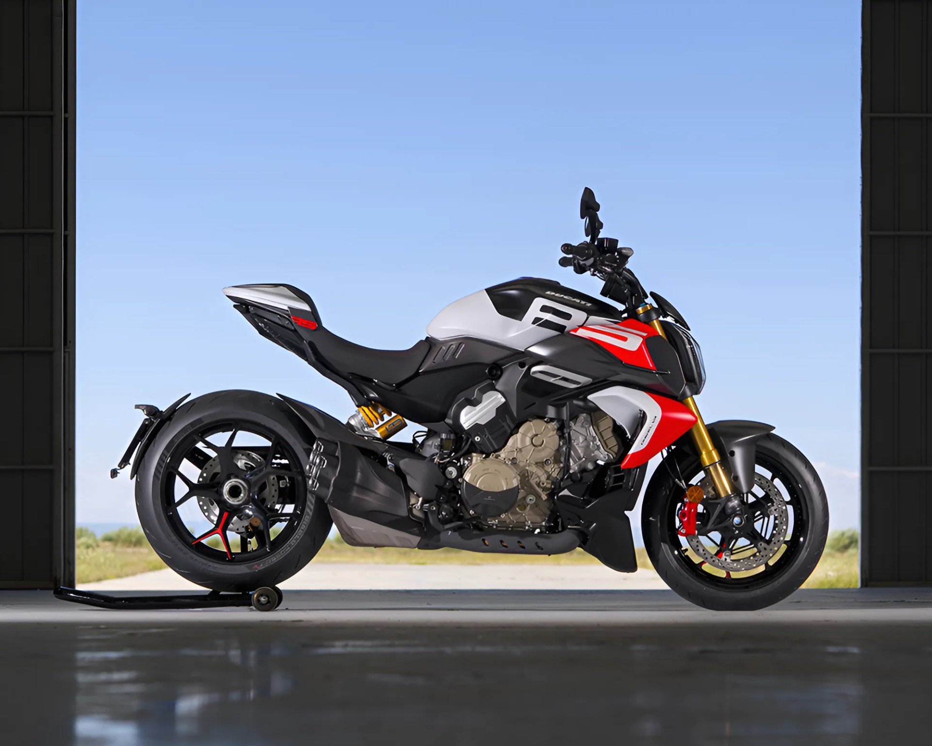 Sport motorcycle with a sleek design, featuring a white, black, and red color scheme. It has a visible engine, gold front forks, and black wheels with red accents. The bike is positioned on a stand inside a garage or hangar, with a clear blue sky and greenery visible outside.