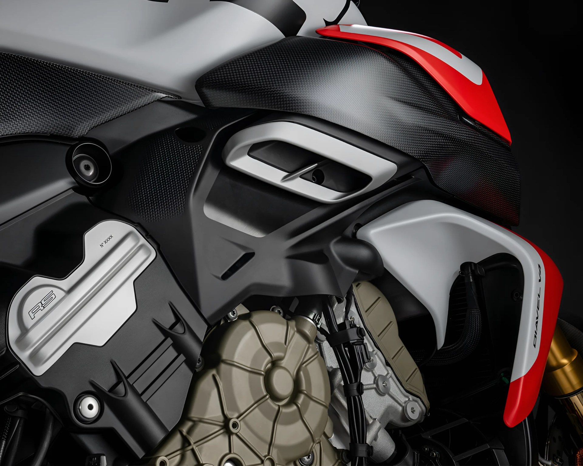 Close-up of a motorcycle engine and surrounding components, featuring a mix of black, silver, and red parts. The engine casing is metallic with a circular design, and the black panel nearby has "RS" and "N° XXXX" markings. The red and white bodywork above the engine includes a carbon fiber texture and the text "DIAVEL V4" on the side.