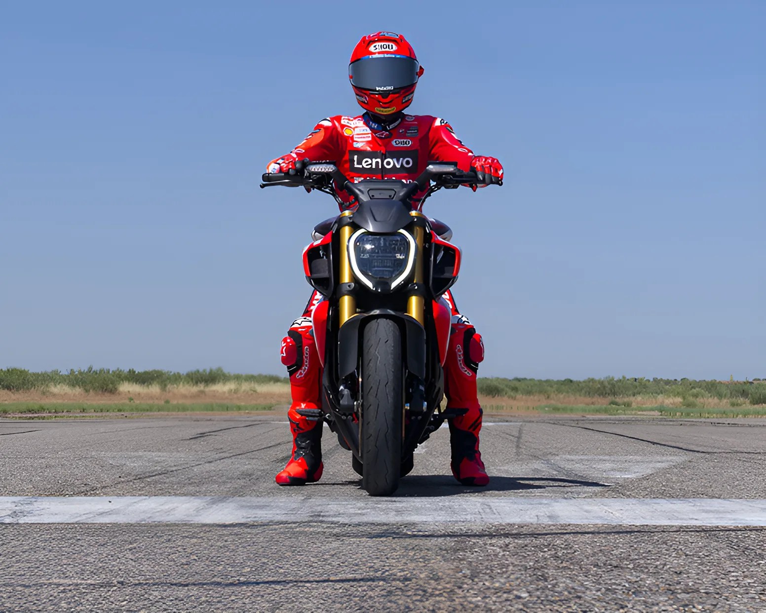 A motorcyclist in a red racing suit and helmet with Lenovo branding is sitting on a black and red motorcycle, positioned on a paved road with a clear blue sky and grassy landscape in the background. The motorcycle is viewed from the front, showing its headlight and wide front tire.