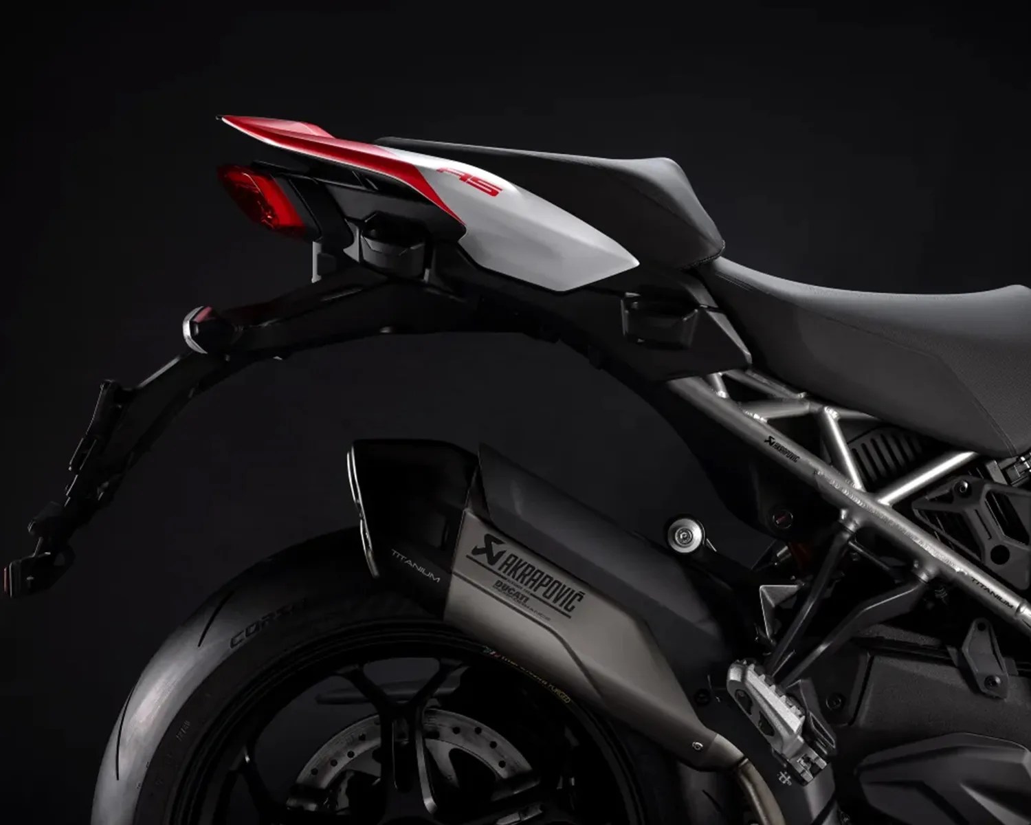 Rear section of a motorcycle featuring a black and silver Akrapovič titanium exhaust, a black rear tire, a red and white tail with a red brake light, and a black seat with a silver frame.