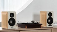 A wooden turntable with copper-colored legs is placed on a light wood surface, flanked by two bookshelf speakers with light wood sides and white front panels. Each speaker has a black tweeter and woofer, with the left speaker labeled "EDIFIER" and the right speaker labeled "CLASSIC." The background features a light gray wall and a white curved architectural detail.