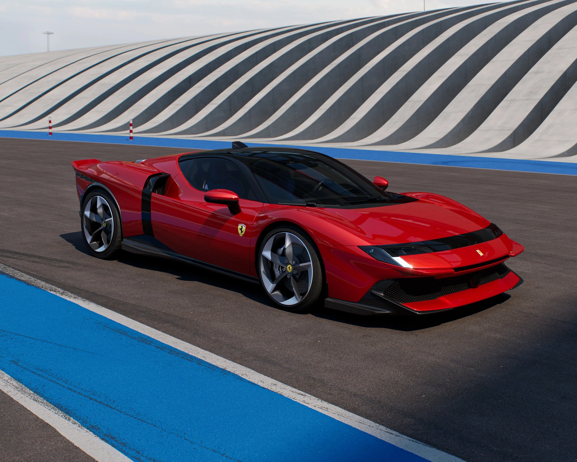 Red Ferrari sports car with sleek, aerodynamic design parked on a racetrack. The car features a low profile, black tinted windows, and distinctive five-spoke alloy wheels. The background shows a curved, striped concrete wall with alternating light and dark gray waves. The racetrack surface is dark asphalt with bright blue and white painted lines along the edge.