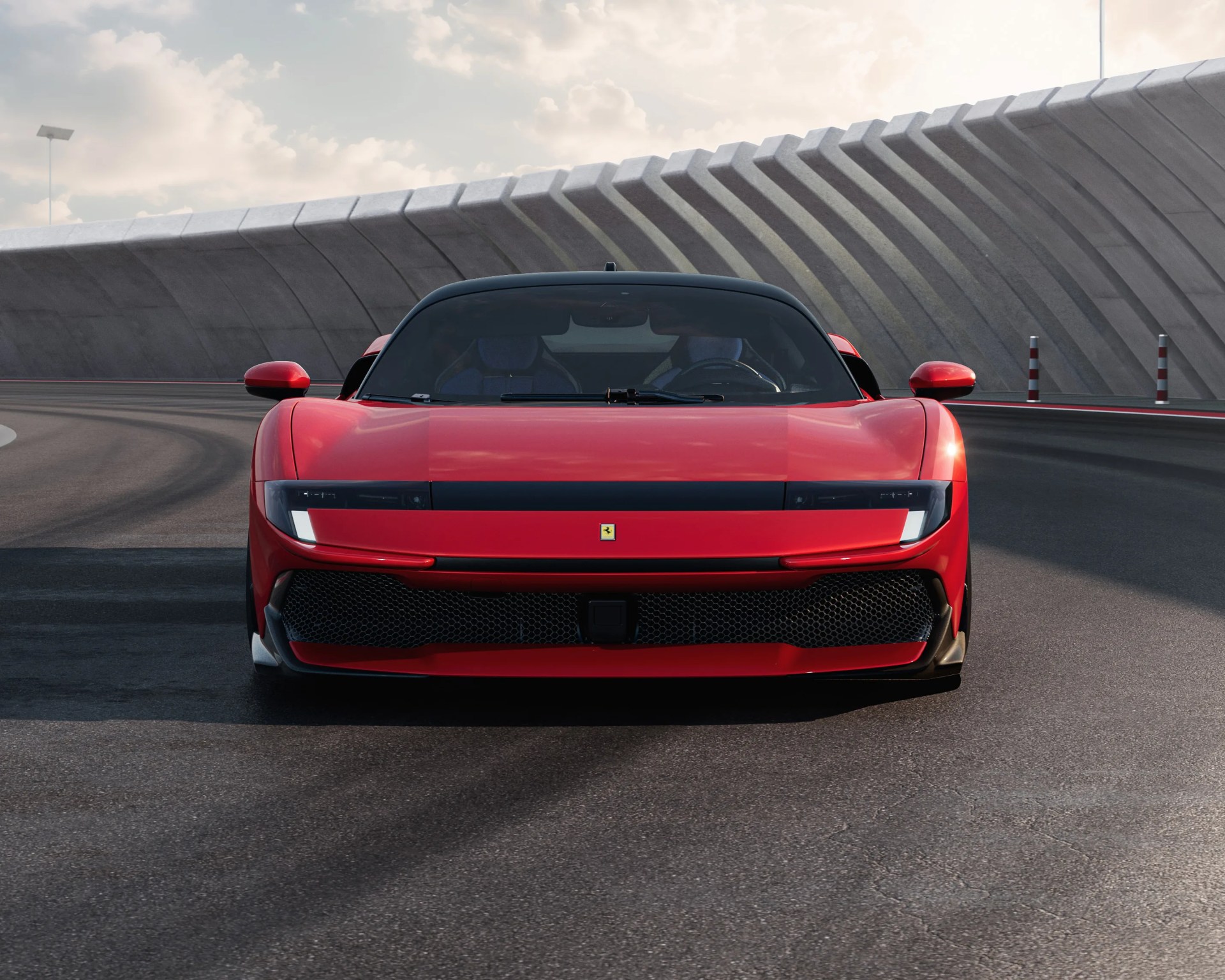 Red Ferrari sports car viewed from the front, positioned on a curved racetrack with a modern concrete barrier in the background. The car features sleek, narrow headlights, a black grille, and a low, aerodynamic profile. The sky is partly cloudy, casting soft light on the scene.