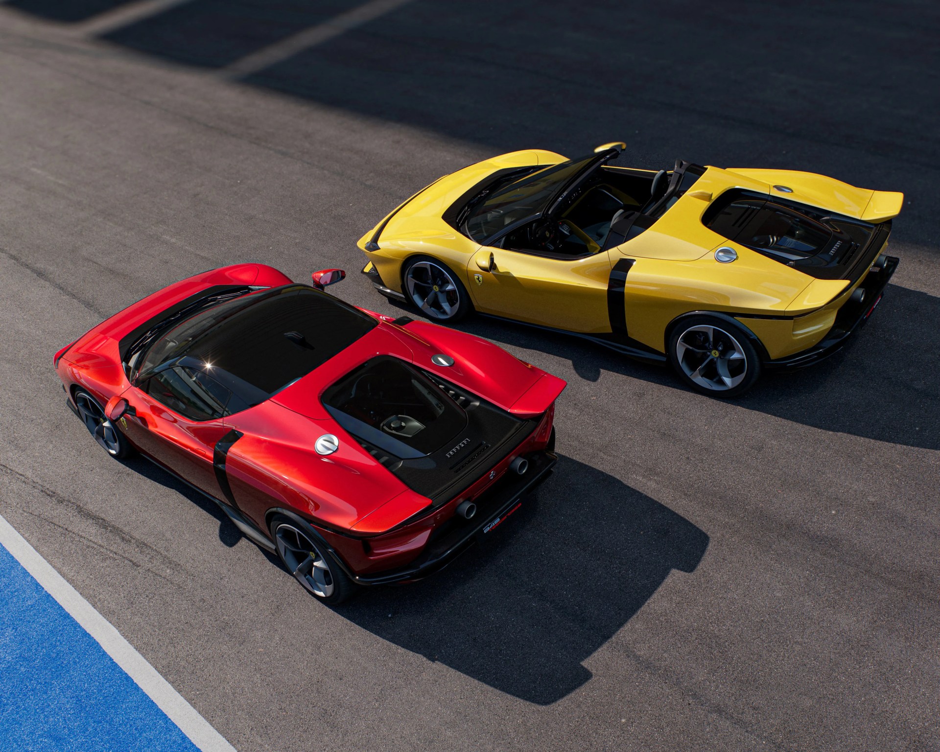 Two Ferrari sports cars parked on a racetrack, one in red and the other in yellow. Both cars have sleek, aerodynamic designs with black accents and visible engine compartments through rear glass panels. The yellow car is a convertible with its roof down, while the red car has a fixed roof. The surface of the racetrack is asphalt with a blue and white painted edge visible in the bottom left corner.