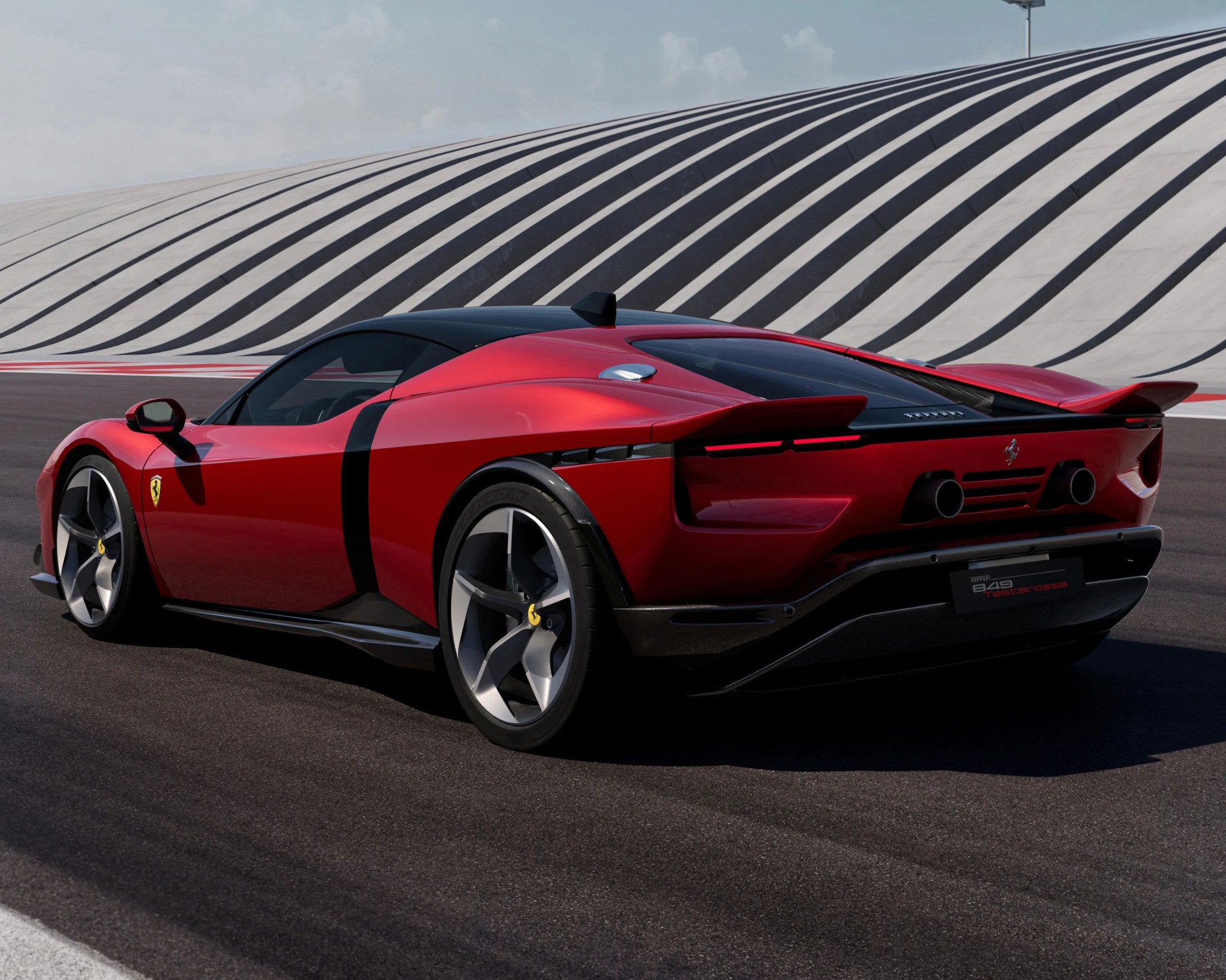 Red Ferrari 849 Tascrossa sports car viewed from the rear side, featuring sleek aerodynamic design, black roof, dual exhausts, and large alloy wheels, parked on a racetrack with a modern, striped architectural backdrop.