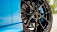 Close-up of a black Ford alloy wheel with a visible blue Brembo brake caliper behind it, mounted on a tire. The car's blue body is partially visible on the left side of the image.