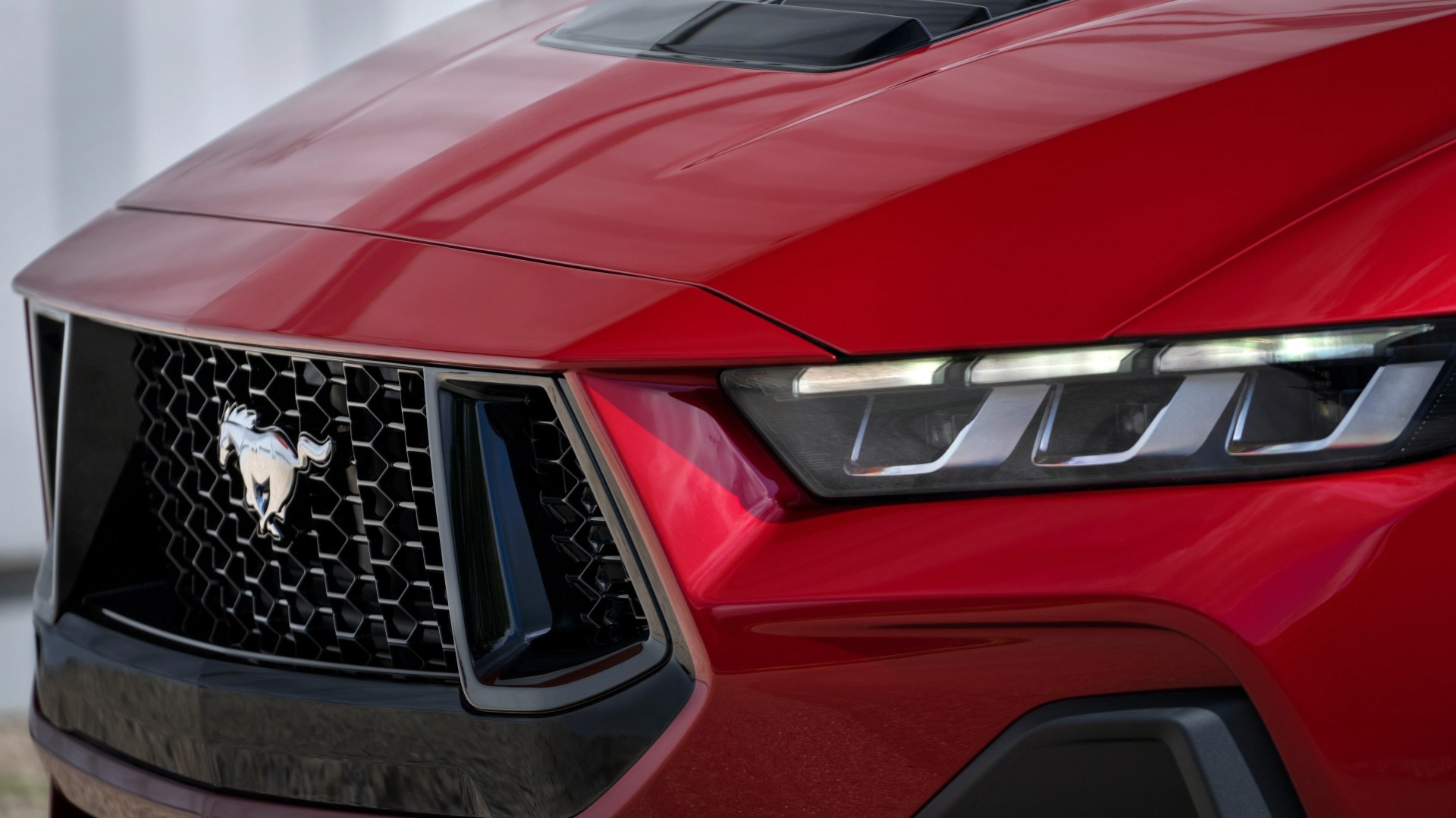 Front view of a red Ford Mustang, focusing on the black honeycomb grille with the iconic silver Mustang horse emblem and the sleek, modern LED headlight on the right side. The hood features a black air vent scoop.