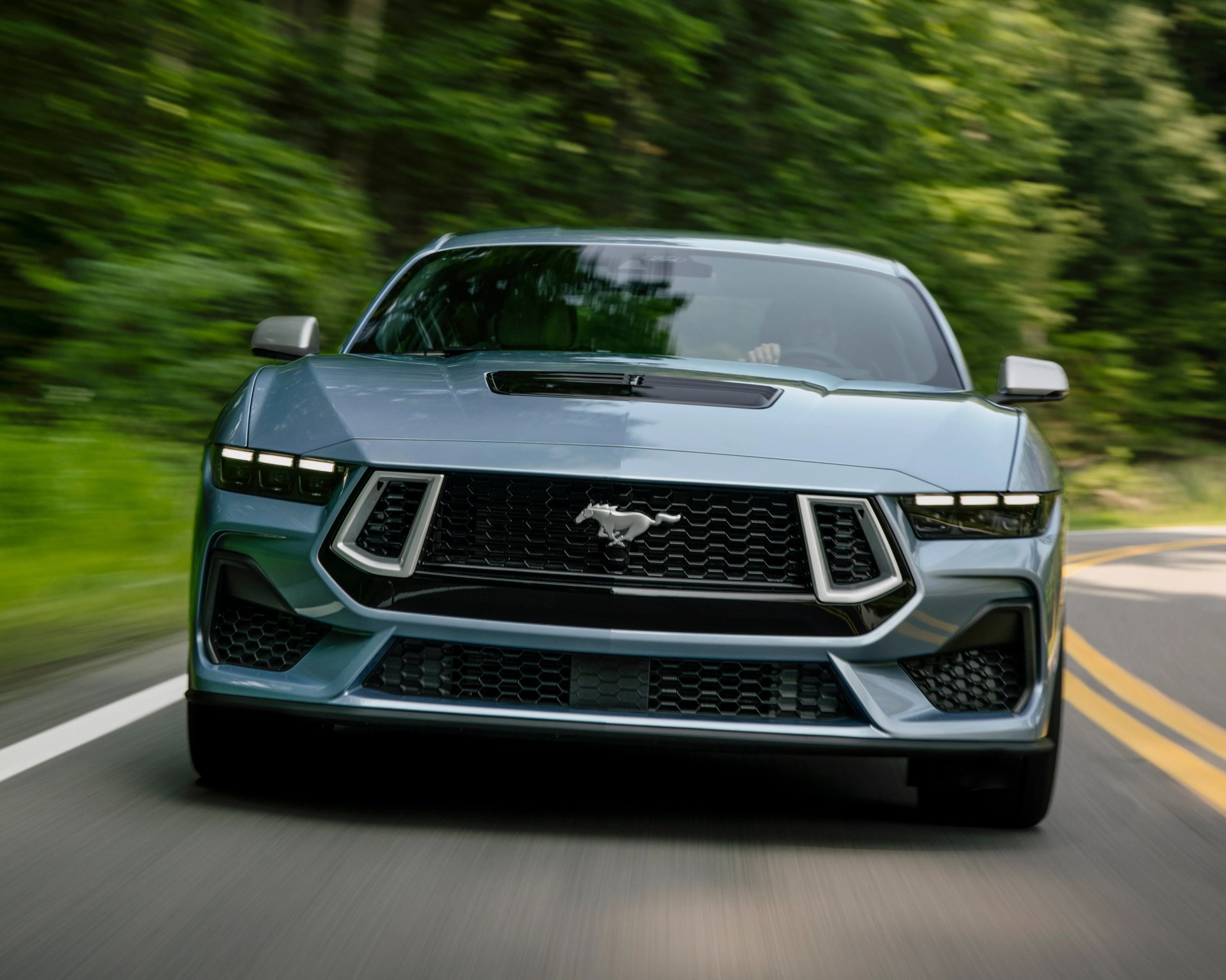Front view of a blue Ford Mustang driving on a winding road with green trees in the background. The car features a prominent black grille with the Mustang emblem, sleek LED headlights, and a hood vent.