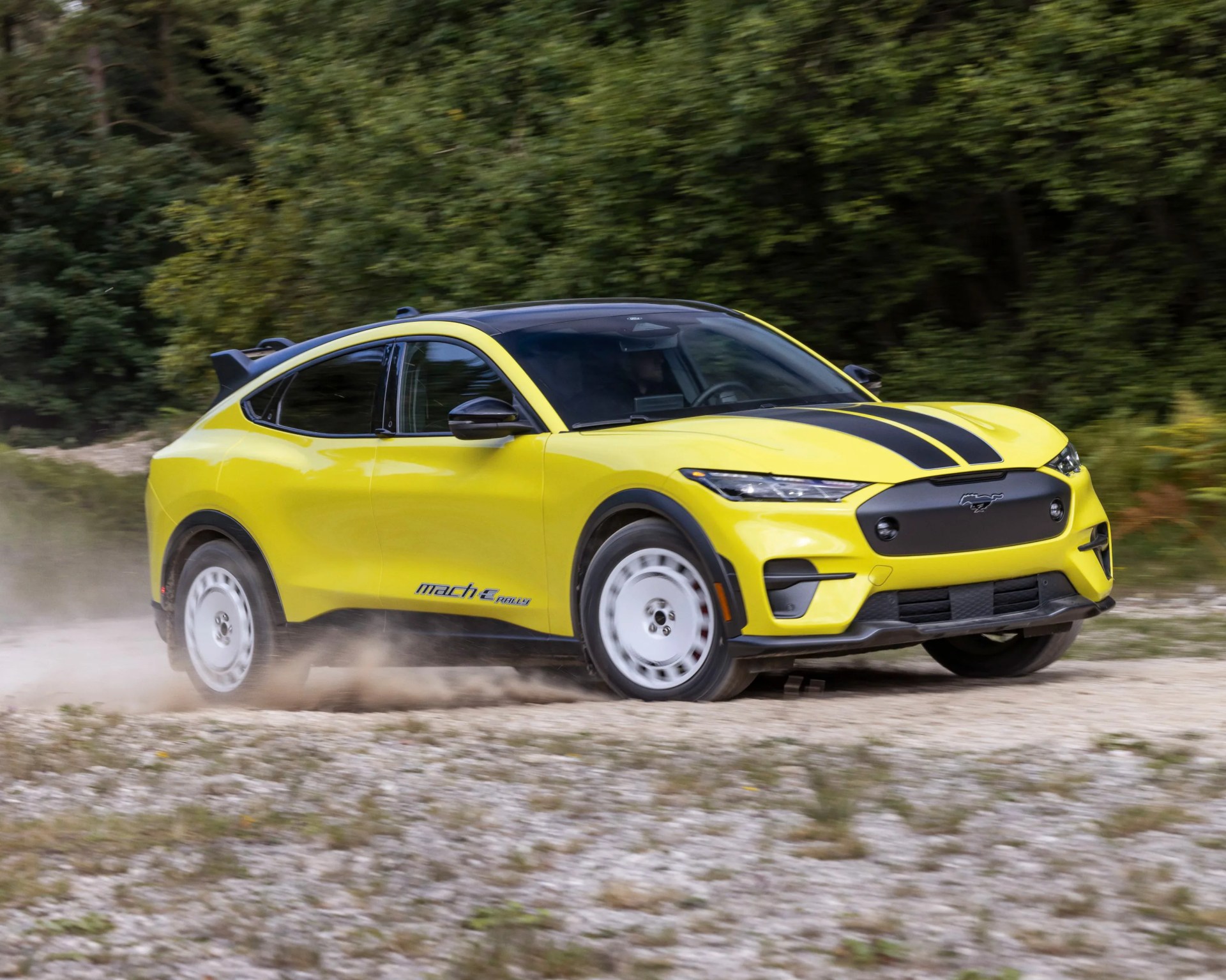 Yellow Ford Mustang Mach-E electric SUV with black racing stripes on the hood, driving on a gravel road with dust kicking up behind the rear wheels, surrounded by green trees.