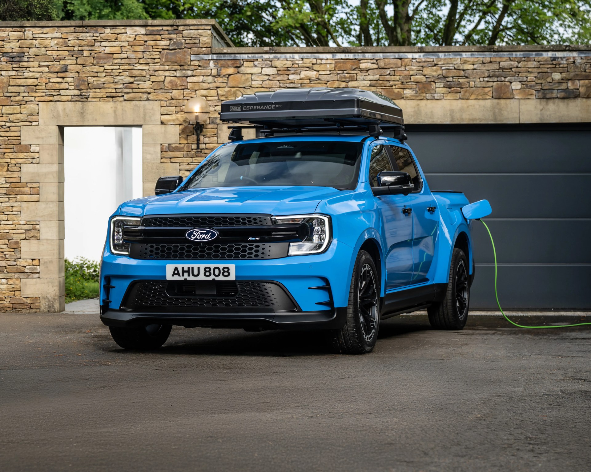 Blue Ford electric pickup truck with a roof-mounted cargo box, parked in front of a stone wall and garage door, connected to a green charging cable.