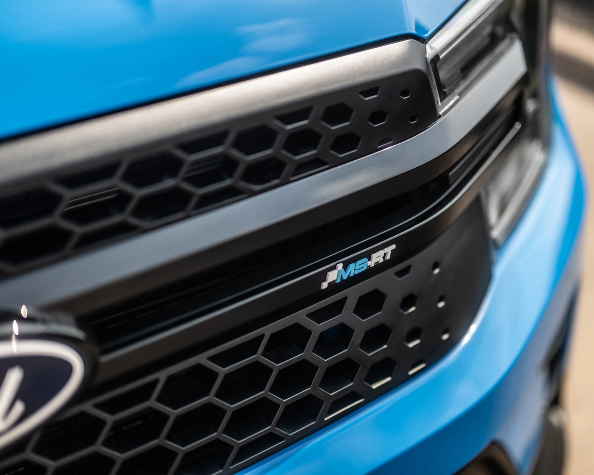 Close-up of a blue car's front grille featuring a black honeycomb pattern and an "MS-RT" badge in blue and white. The grille is bordered by black trim, and part of the car's headlight and emblem are visible.