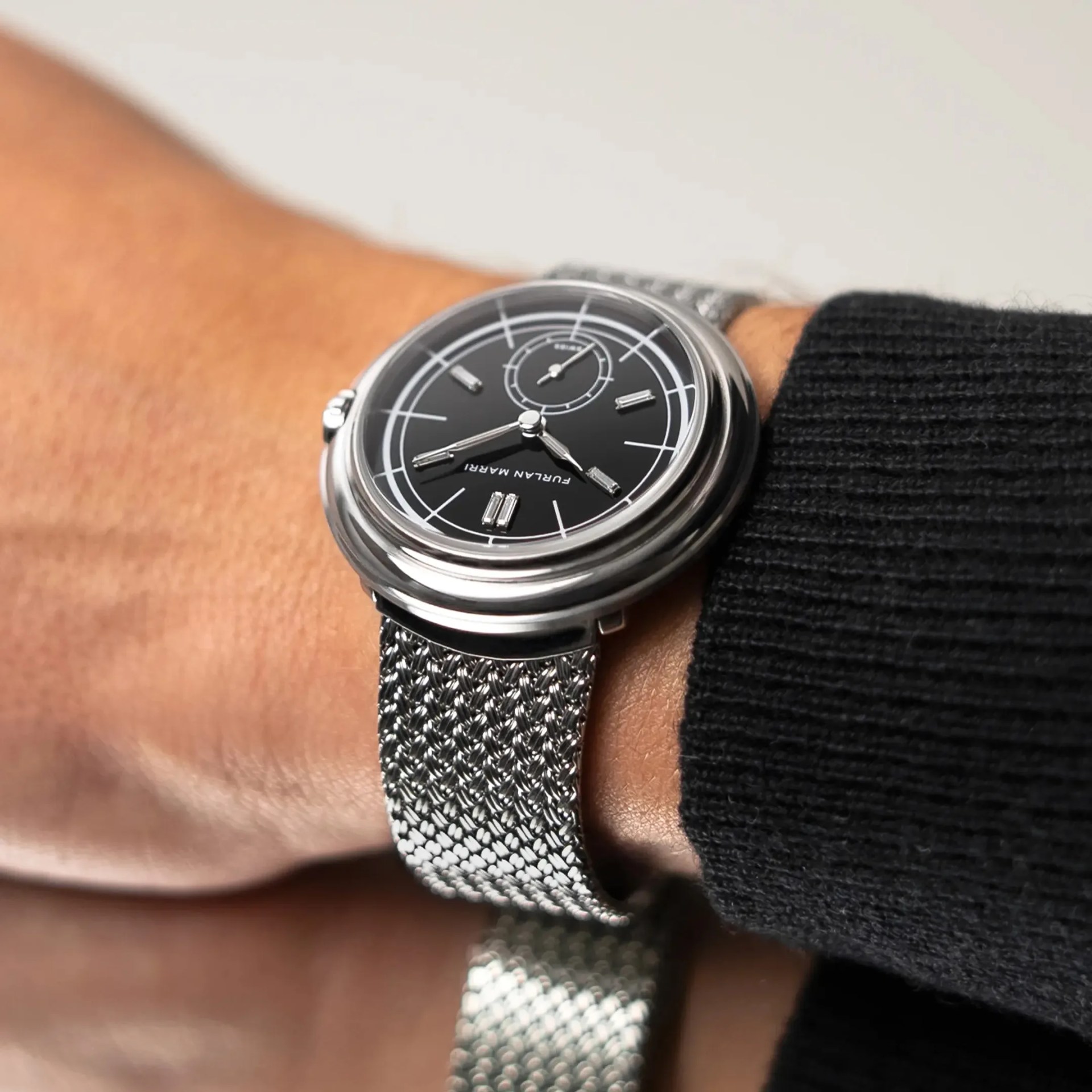 Silver wristwatch with a black dial featuring white markings and the text "FURLAN MARRI," worn on a wrist with a silver mesh strap and a black ribbed sleeve.