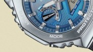 Close-up of a silver wristwatch with a blue dial, showing part of the bezel engraved with the words "ADJUST," "MODE," and "PROTECTION." The watch face features silver hour markers and hands, a small sub-dial with black and white indicators, and additional text including "RCVD REM HND." The watch has a textured metallic finish and a rugged, sporty design.