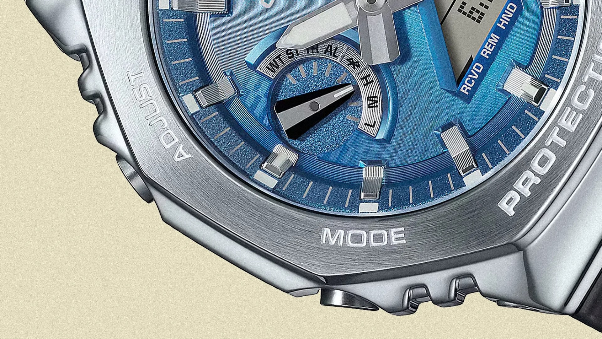 Close-up of a silver wristwatch with a blue dial, showing part of the bezel engraved with the words "ADJUST," "MODE," and "PROTECTION." The watch face features silver hour markers and hands, a small sub-dial with black and white indicators, and additional text including "RCVD REM HND." The watch has a textured metallic finish and a rugged, sporty design.