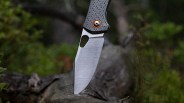 A close-up of a folding knife with a textured handle featuring a geometric pattern and an orange ring around the pivot screw. The blade is partially serrated and inserted into a piece of wood outdoors, with blurred greenery in the background.