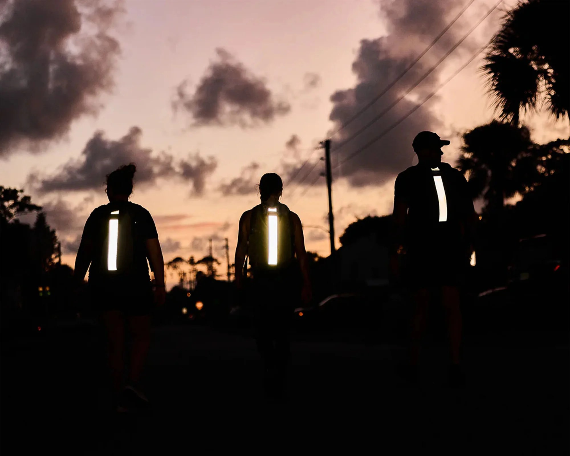 Three people walking outdoors at dusk or dawn, each wearing a backpack with a vertical reflective strip that is brightly illuminated against the dark silhouettes and cloudy sky.