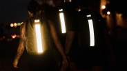 Three people walking at night wearing backpacks with bright reflective vertical strips, with one person on the left showing a tattooed arm.
